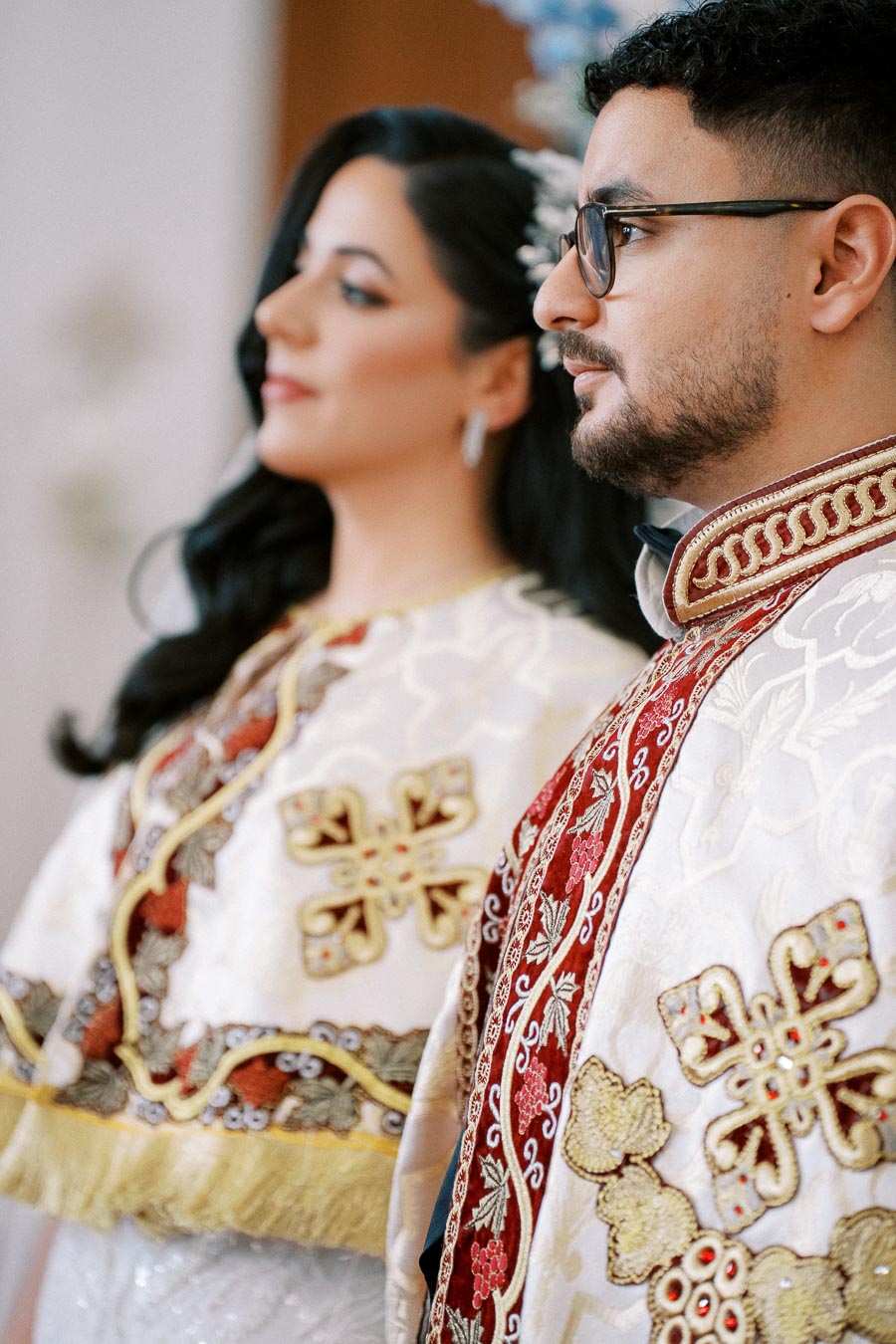 A couple in traditional ornate attire attends a cultural ceremony, showcasing intricate embroidered patterns and vibrant colors.