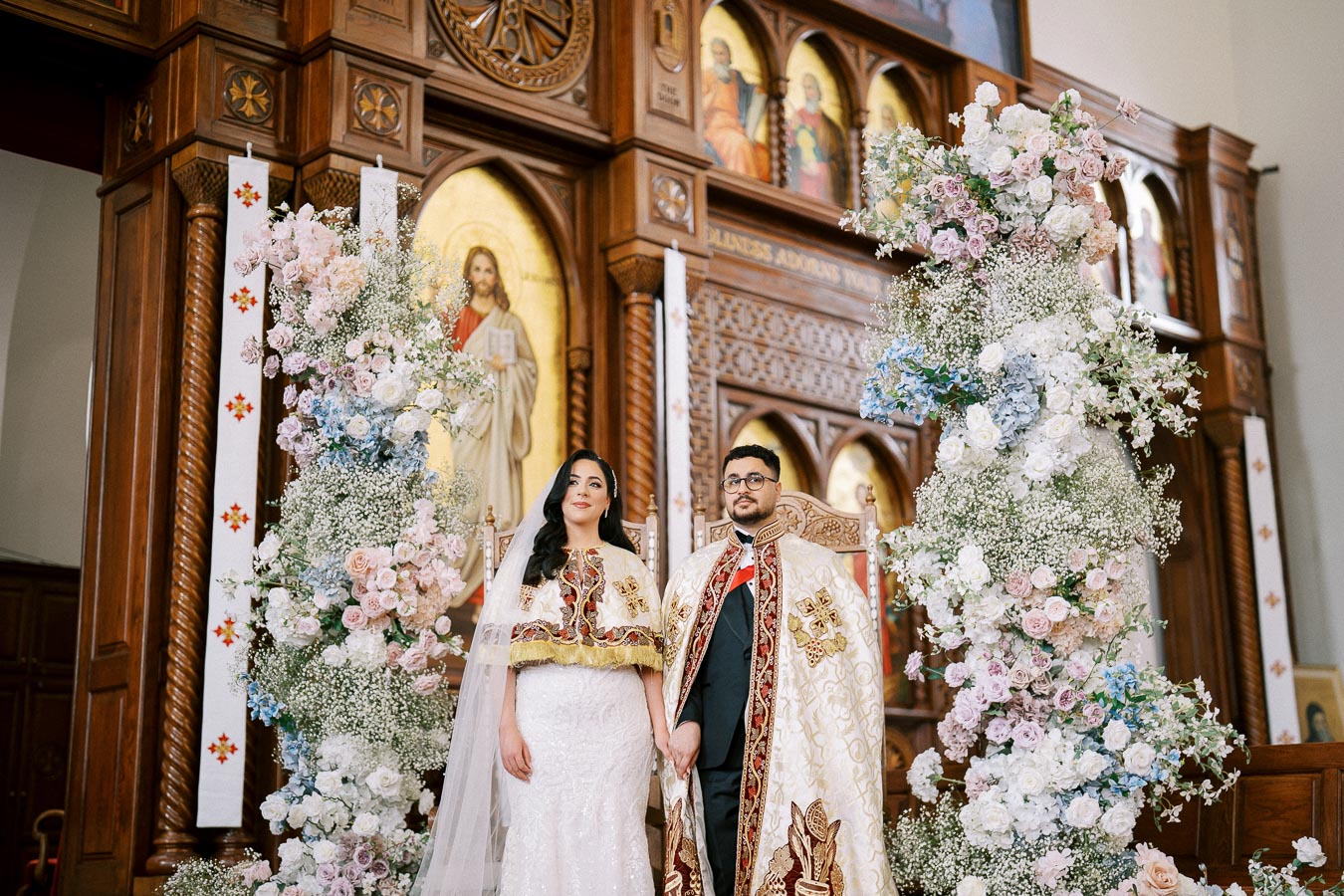 A bride and groom in traditional ceremonial attire stand in an ornate church, surrounded by large floral arrangements and intricate woodwork, during a religious wedding ceremony.