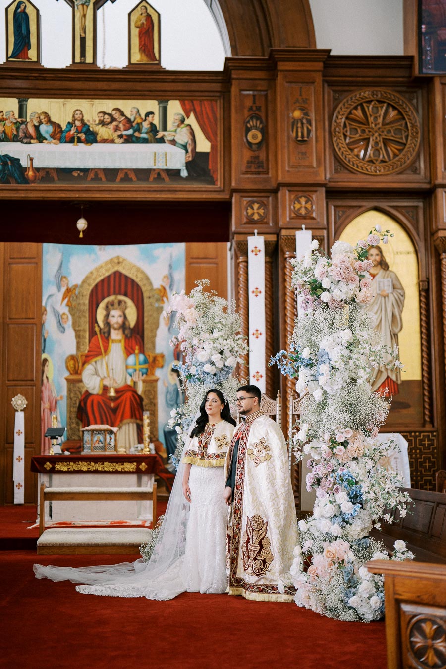 A couple in ornate traditional attire stands inside a beautifully decorated church, with elaborate floral arrangements and religious iconography in the background, capturing the essence of a ceremonial wedding setting.