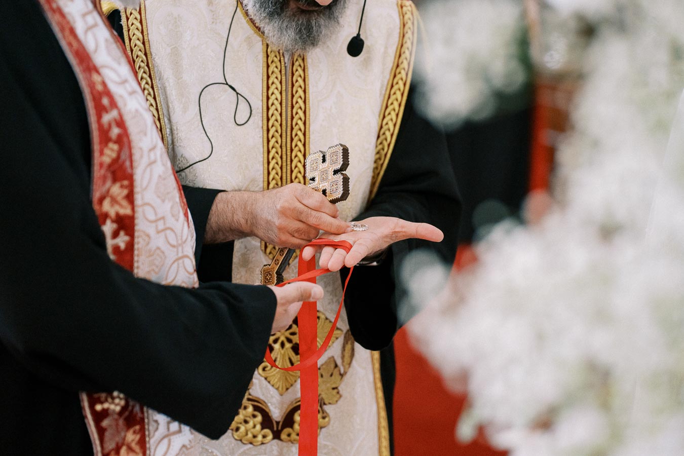 Priest in ornate religious attire performing a ceremonial blessing with a cross, holding a red ribbon.