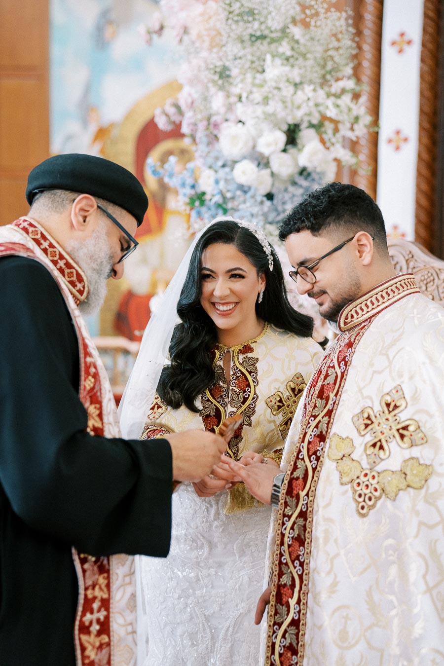 A joyful couple dressed in ornate traditional attire exchanges rings during a wedding ceremony, officiated by a priest in a richly decorated religious setting.