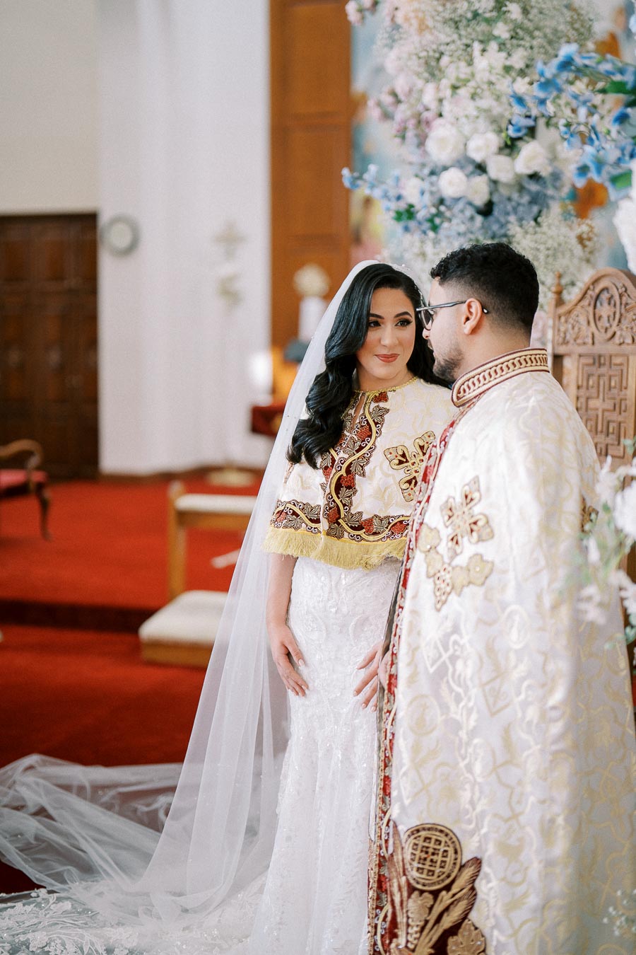 Bride and groom in traditional ornate wedding attire during a ceremonial moment in a beautifully decorated venue with vibrant floral arrangements.
