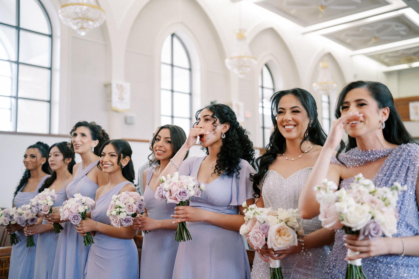 Bridesmaids in light lavender dresses holding pastel bouquets, smiling and standing in a beautifully decorated church, celebrating a wedding ceremony.
