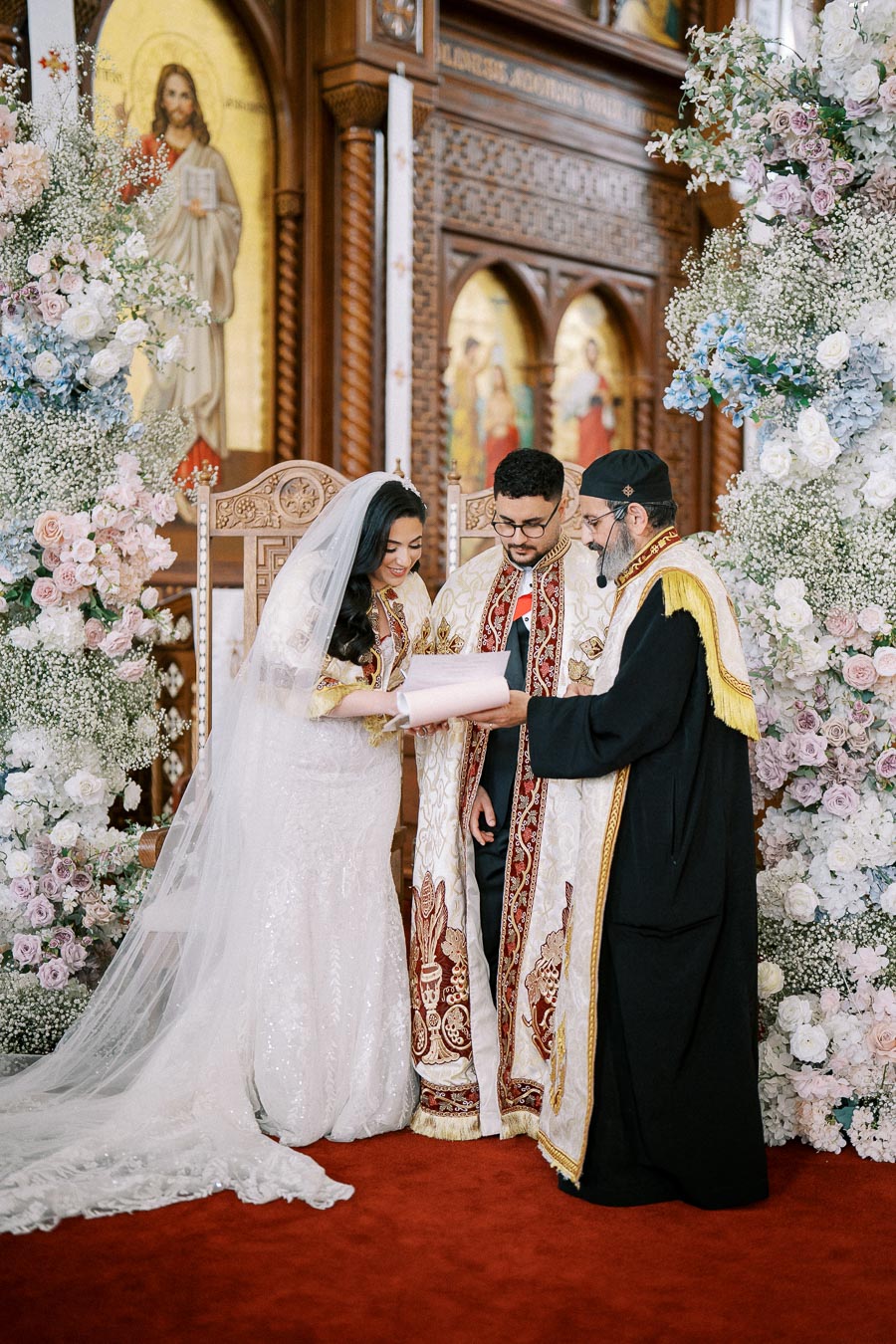 Bride and groom in traditional ceremonial attire stand with a priest in an ornate church, exchanging vows surrounded by elegant floral arrangements.
