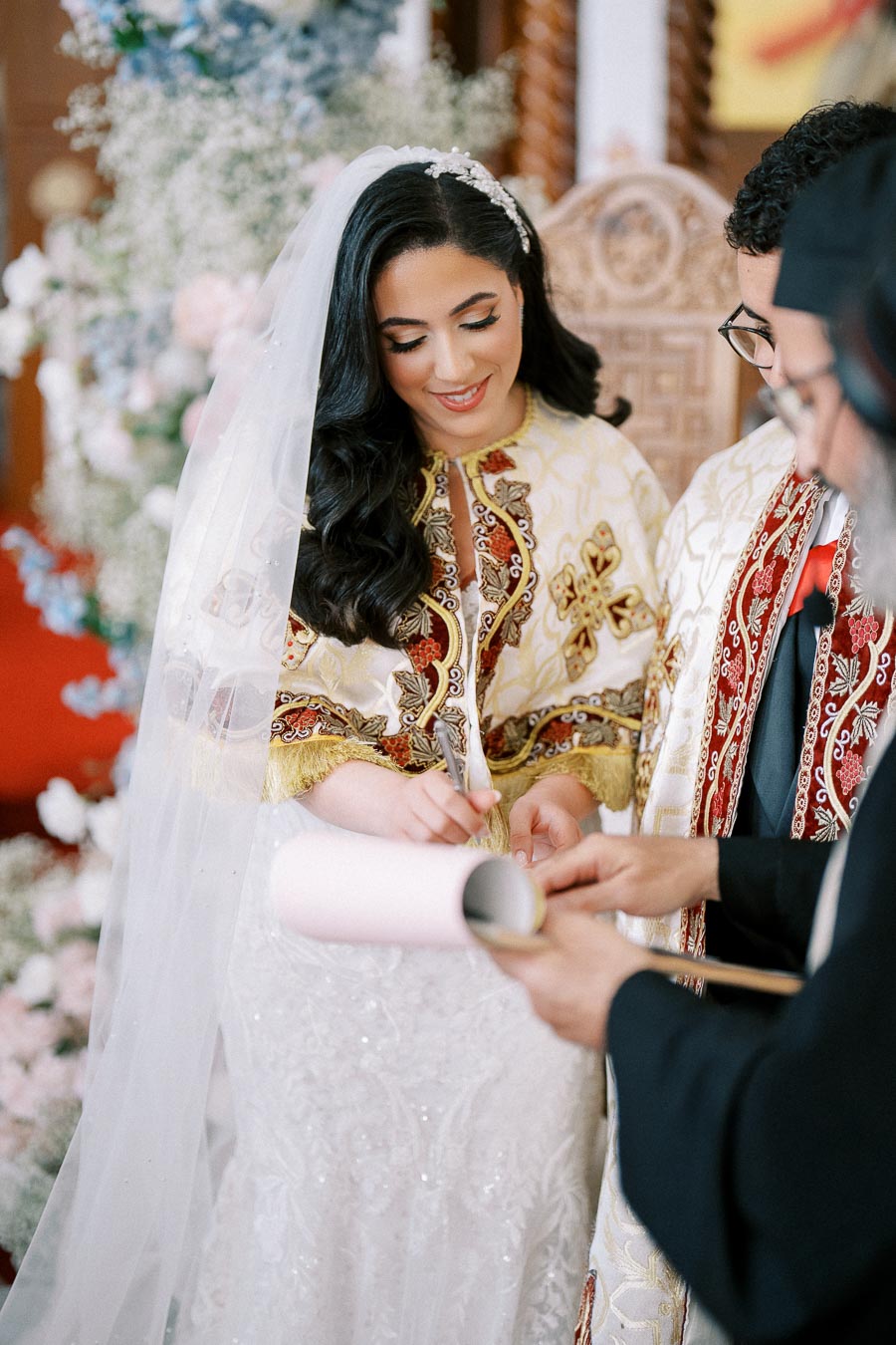 Bride signing wedding documents during a traditional ceremony, wearing an ornate embroidered gown and veil, surrounded by floral decorations.