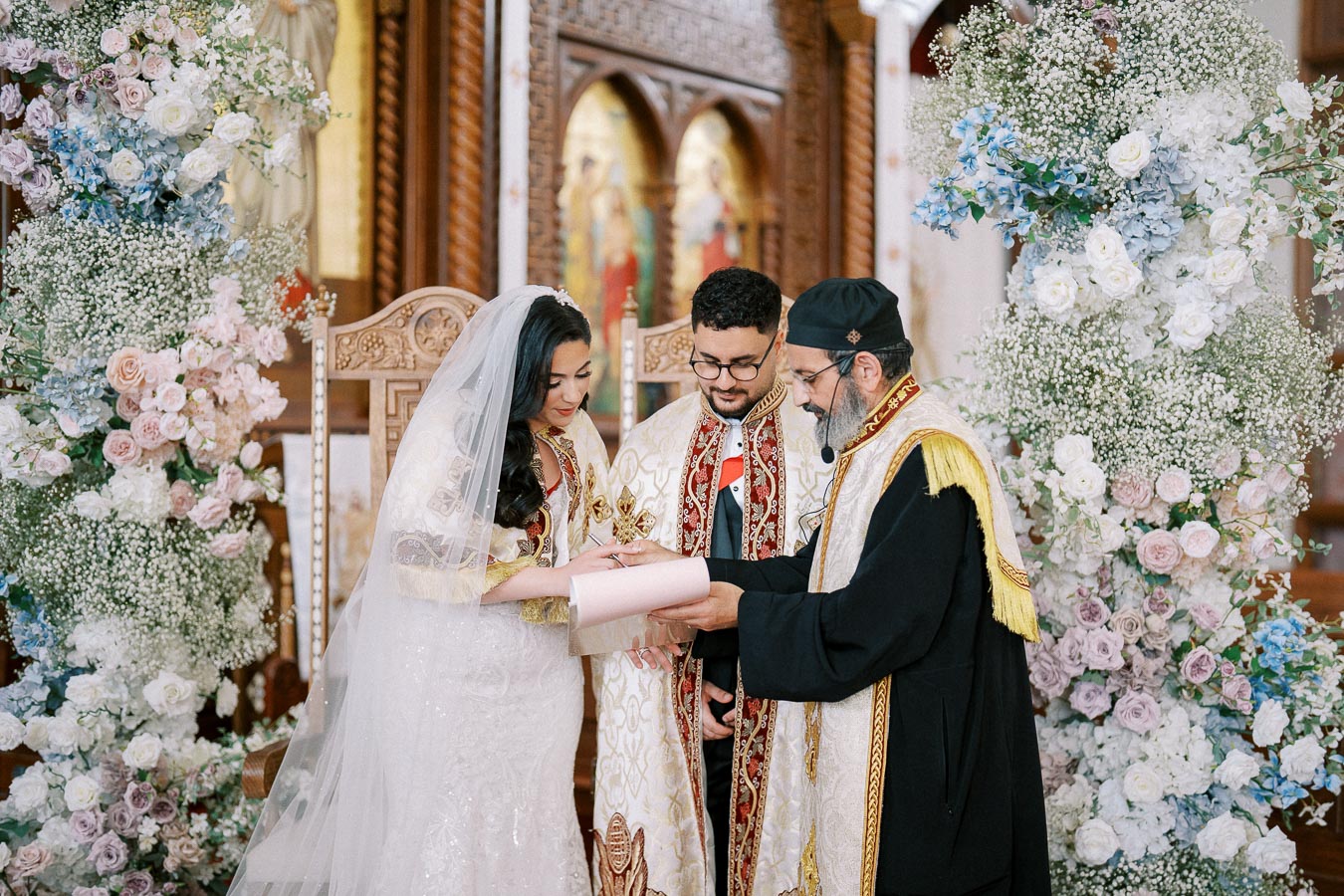 A bride and groom in traditional clothing participate in a wedding ceremony officiated by a priest, surrounded by floral decorations in a church setting.