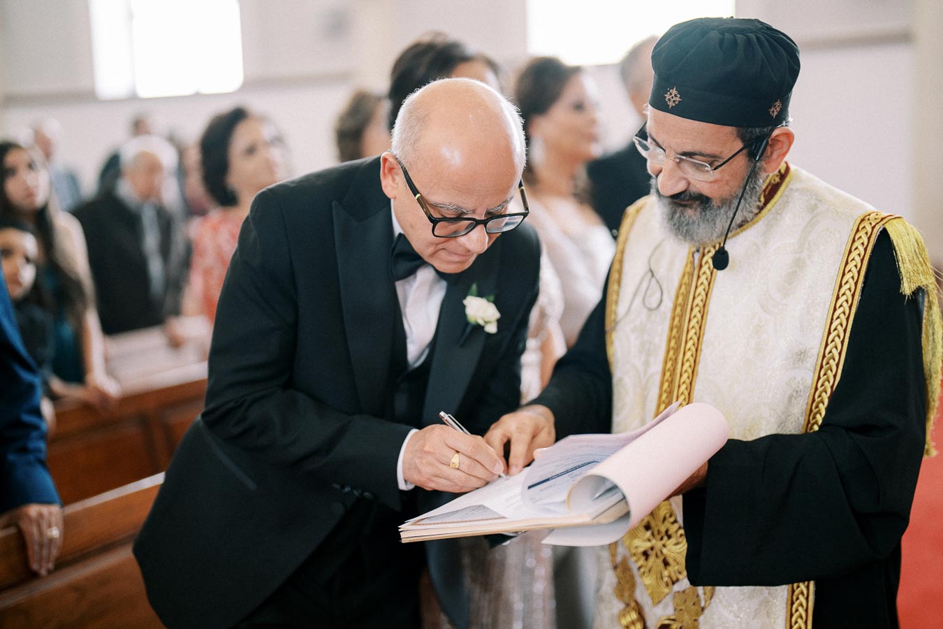 An older man in a formal suit signs a document with a priest in traditional religious attire overseeing the process inside a church setting.