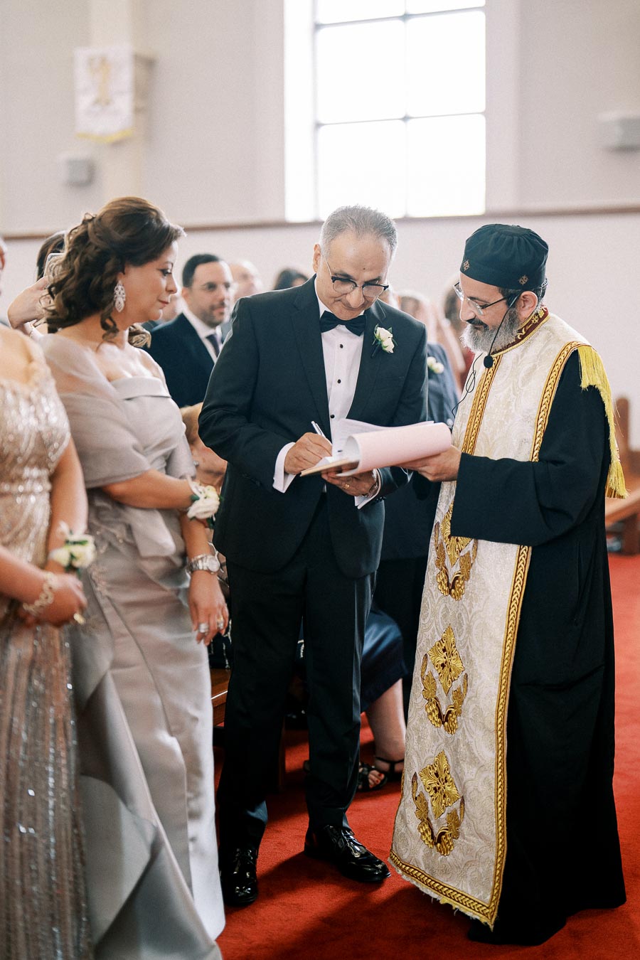 A formal ceremony inside a church, featuring a man in a tuxedo signing a document with a priest in a traditional robe, while elegantly dressed guests look on.
