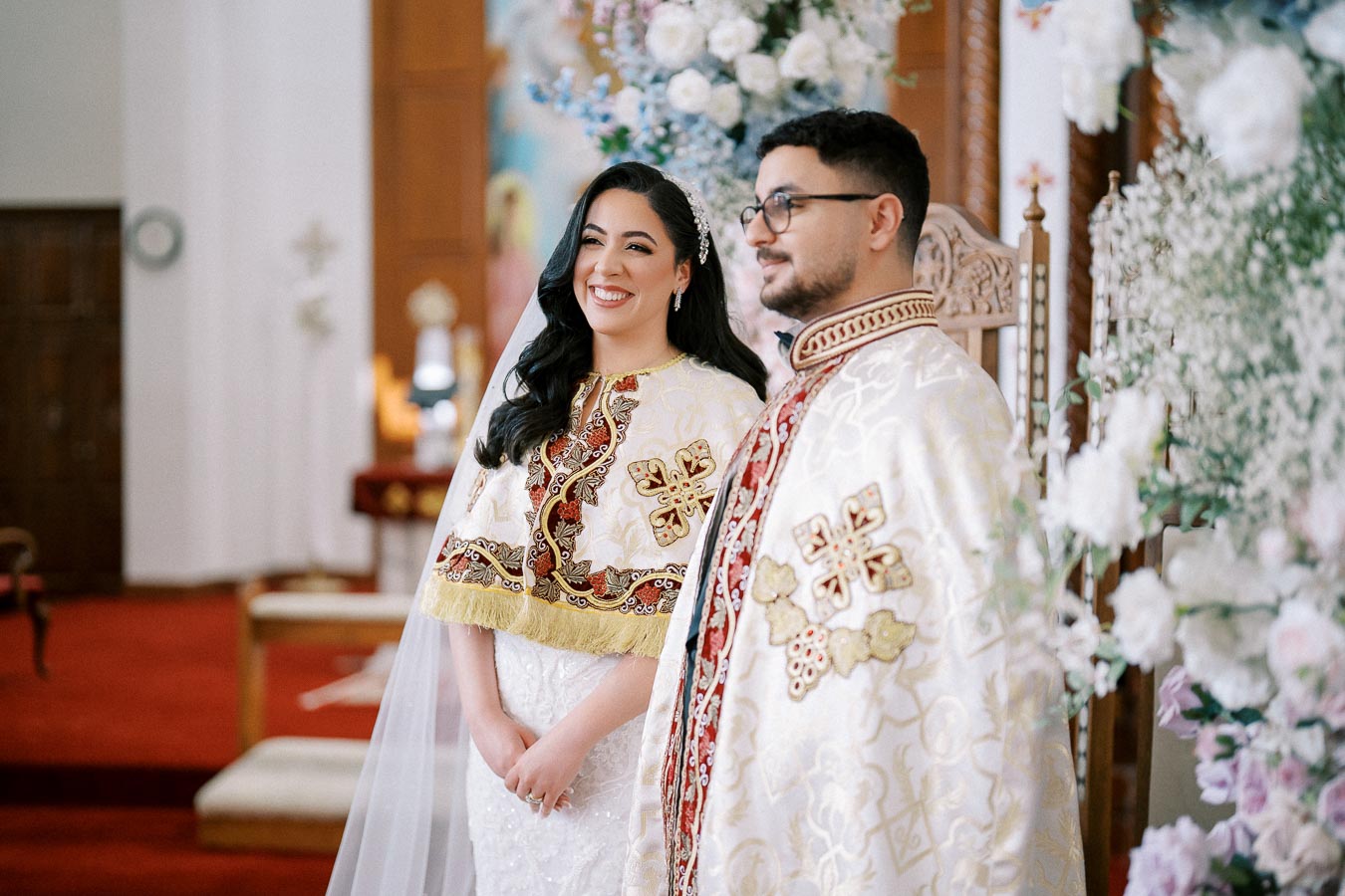 Smiling couple in traditional ceremonial attire with intricate gold and red embroidery, standing in a beautifully decorated church filled with flowers and vibrant colors.