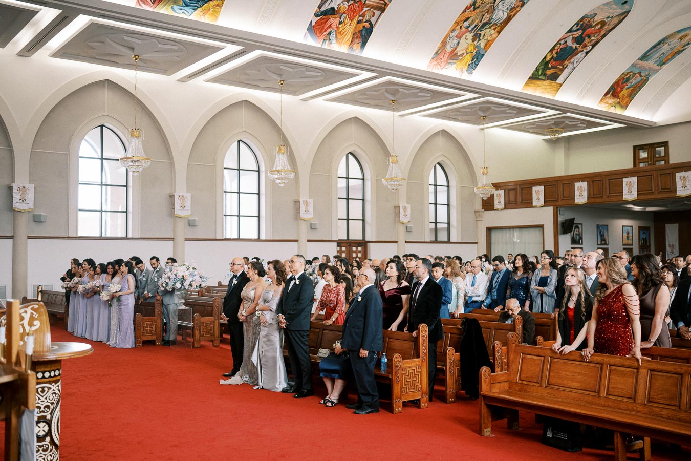A wedding ceremony in a beautifully decorated church, featuring elegantly dressed guests and a bridal party with pastel floral arrangements. The interior showcases arched windows, traditional wooden pews, and colorful ceiling artwork, creating a serene and festive atmosphere.