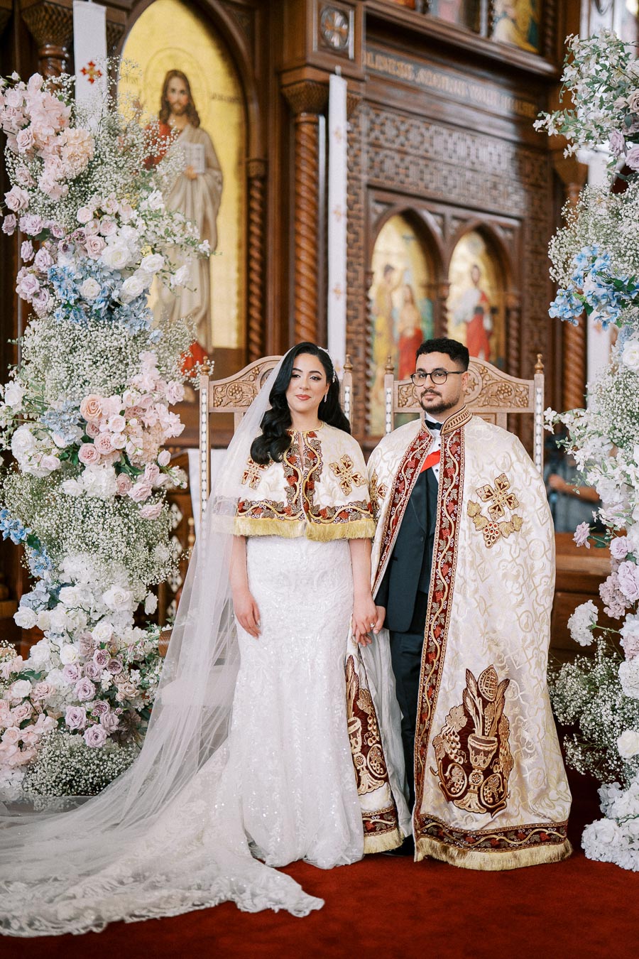 A couple dressed in ornate traditional wedding attire stands hand in hand inside a beautifully decorated church with floral arrangements and religious icons.