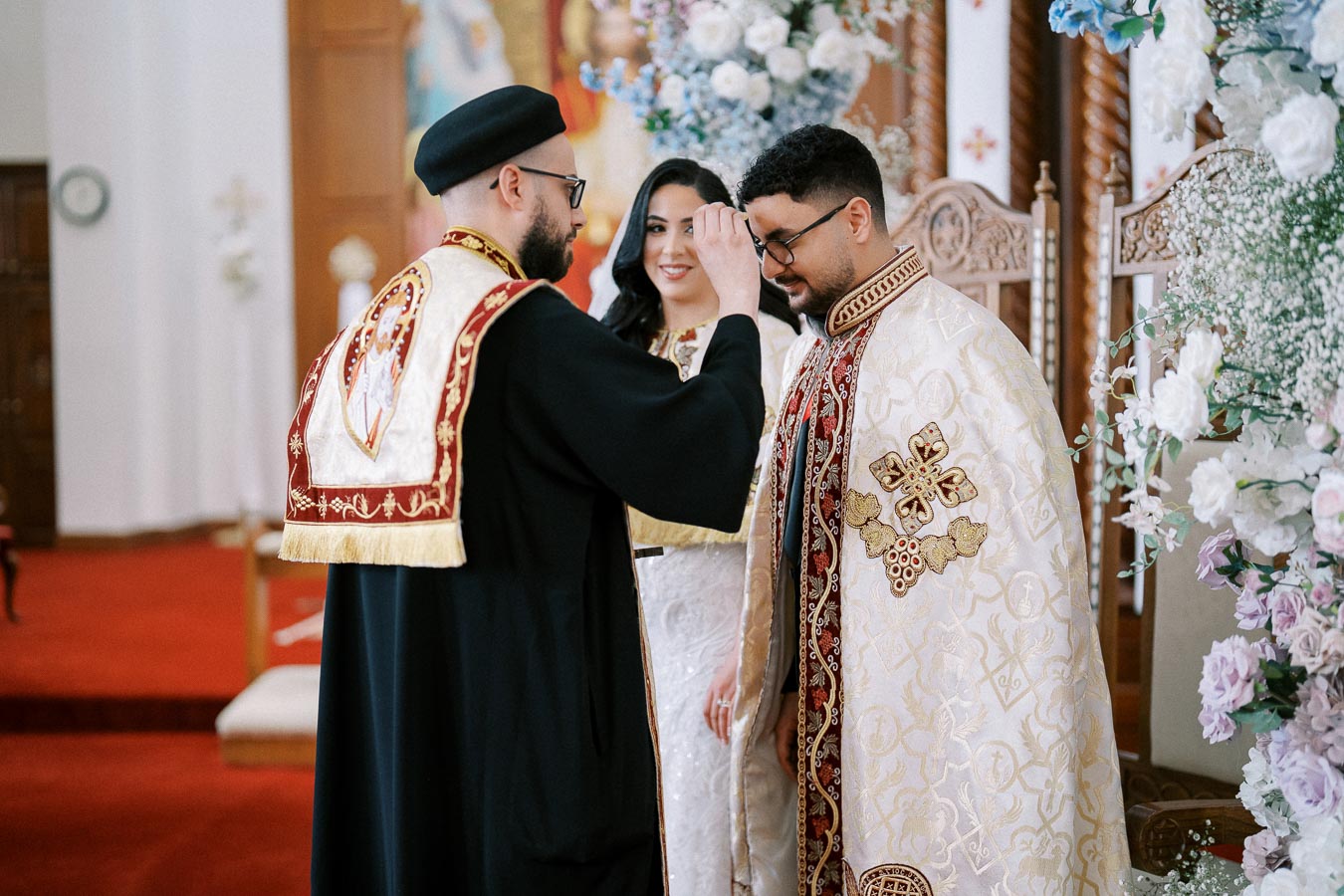 Cleric performing a traditional religious blessing inside a decorated church, with an ornately dressed couple standing nearby, surrounded by floral arrangements.