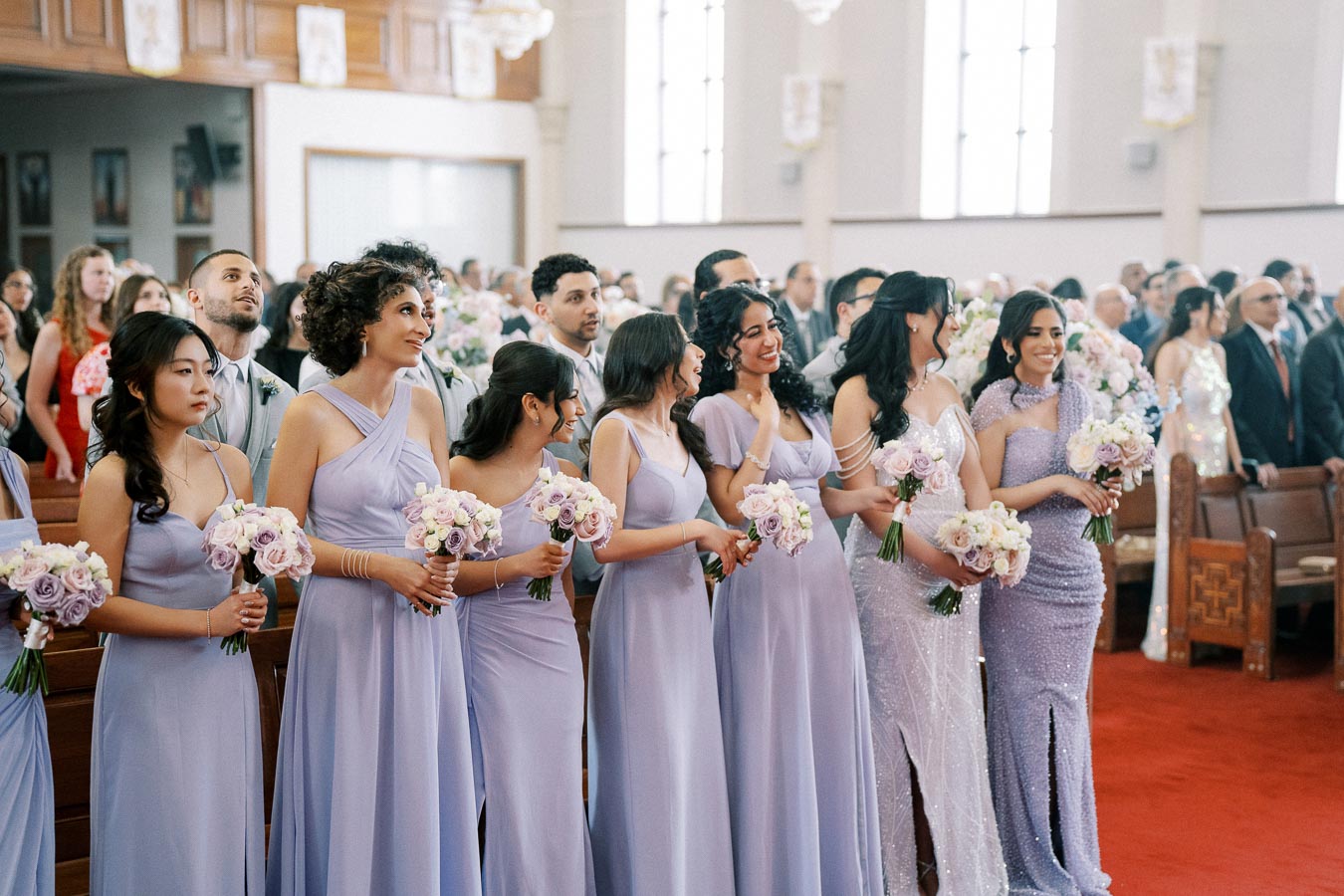 Bridesmaids in lavender dresses holding pastel bouquets in a church setting, surrounded by guests during a wedding ceremony.