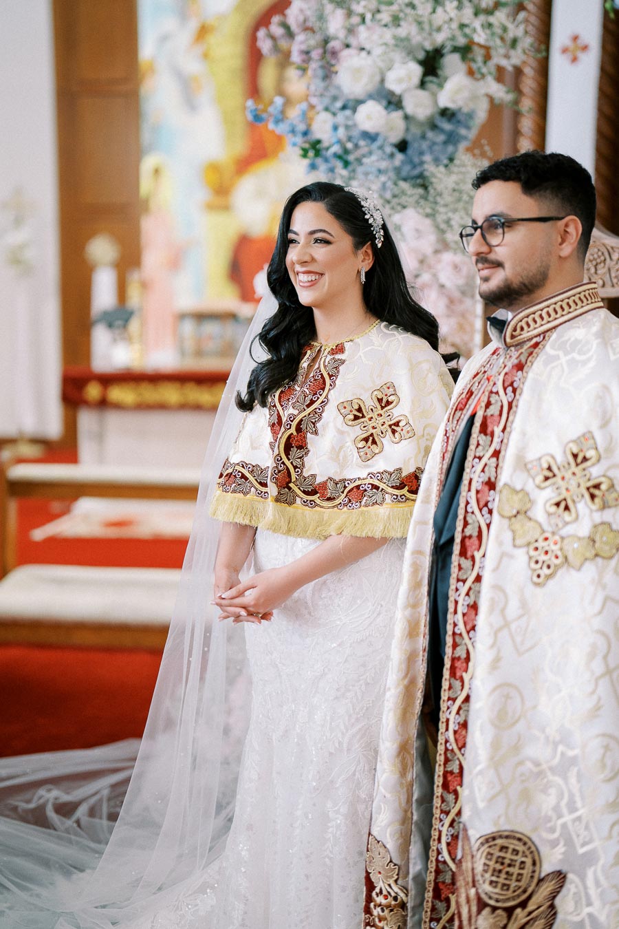 A bride and groom in ornate white and red traditional attire during a wedding ceremony at a beautifully decorated church.