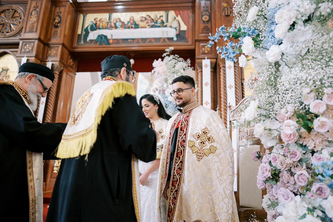 A bride and groom stand before a priest in an ornate church ceremony, surrounded by elaborate floral arrangements and religious iconography.