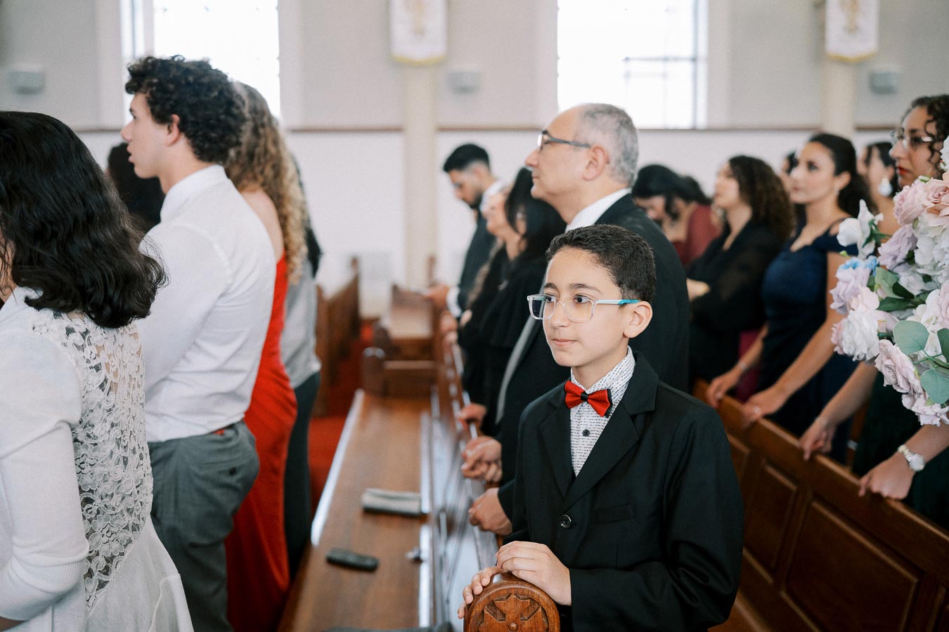 A group of people in formal attire attending a church service, with a young boy in a black suit and red bowtie standing in the foreground, highlighting a solemn and respectful atmosphere.
