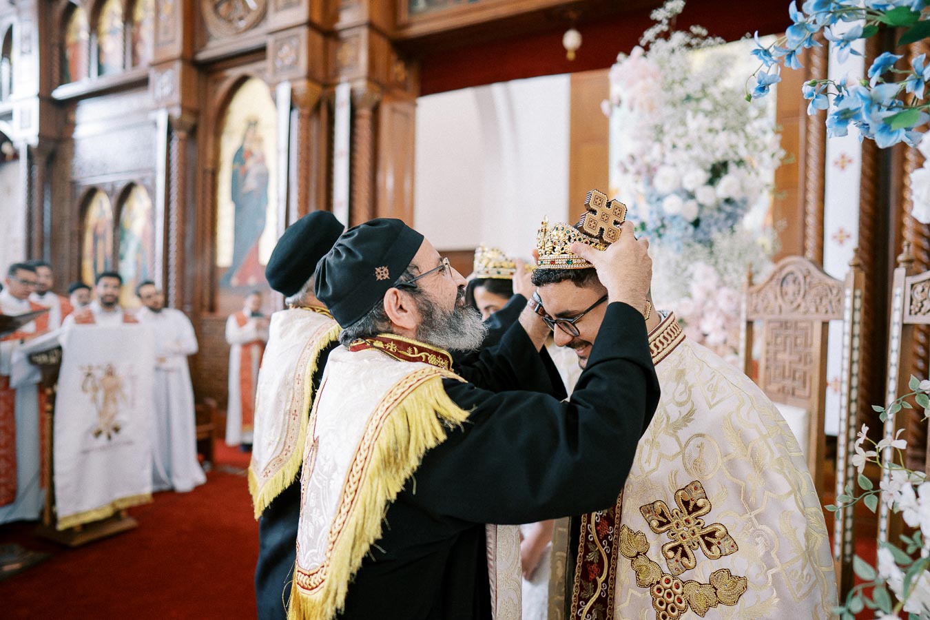 Orthodox priest places a ceremonial crown on a bridegroom during a traditional church wedding ceremony, surrounded by ornate religious decorations and fellow clergy members.