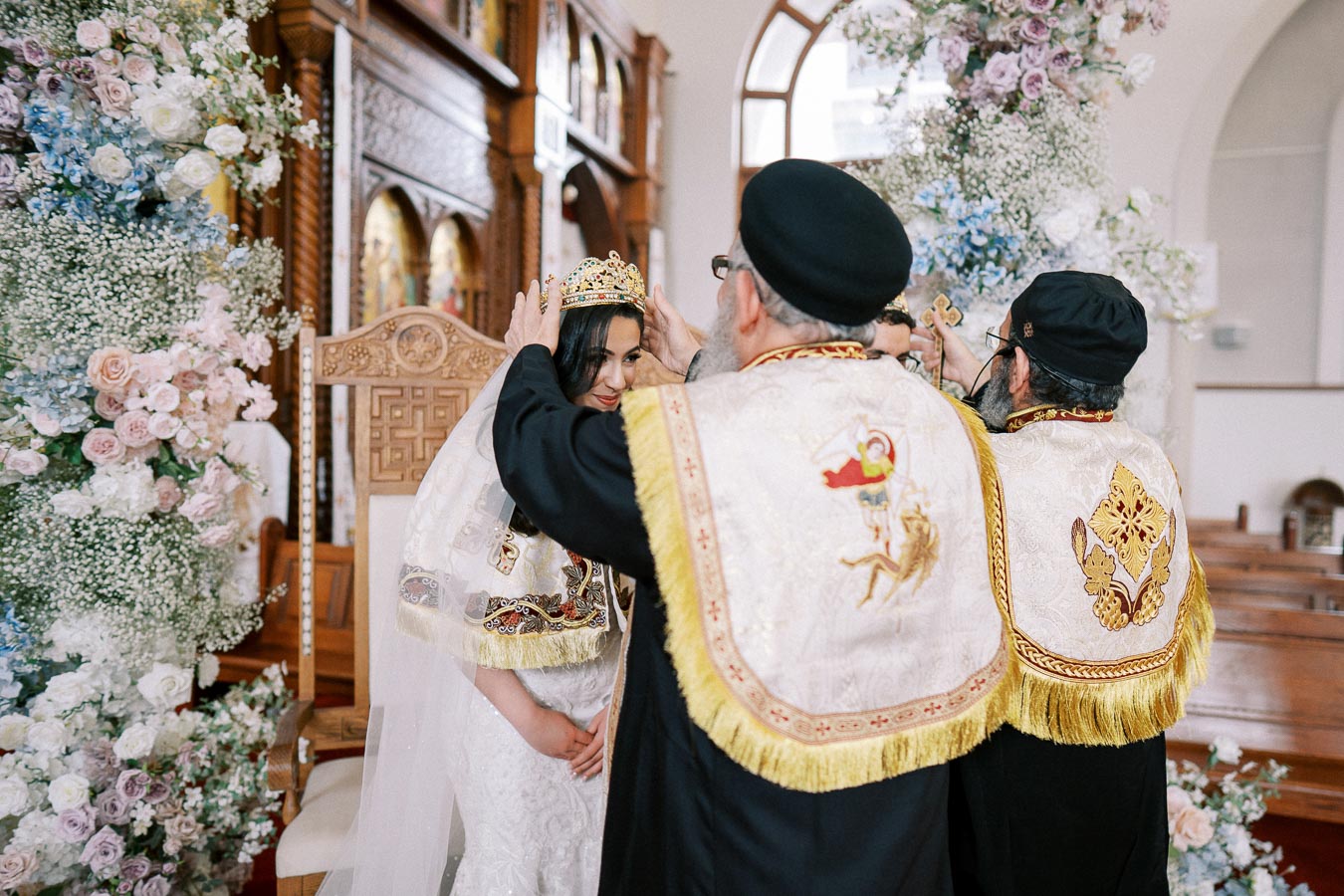 A religious ceremony in a beautifully decorated church, featuring a priest placing a crown on a bride, surrounded by floral arrangements.