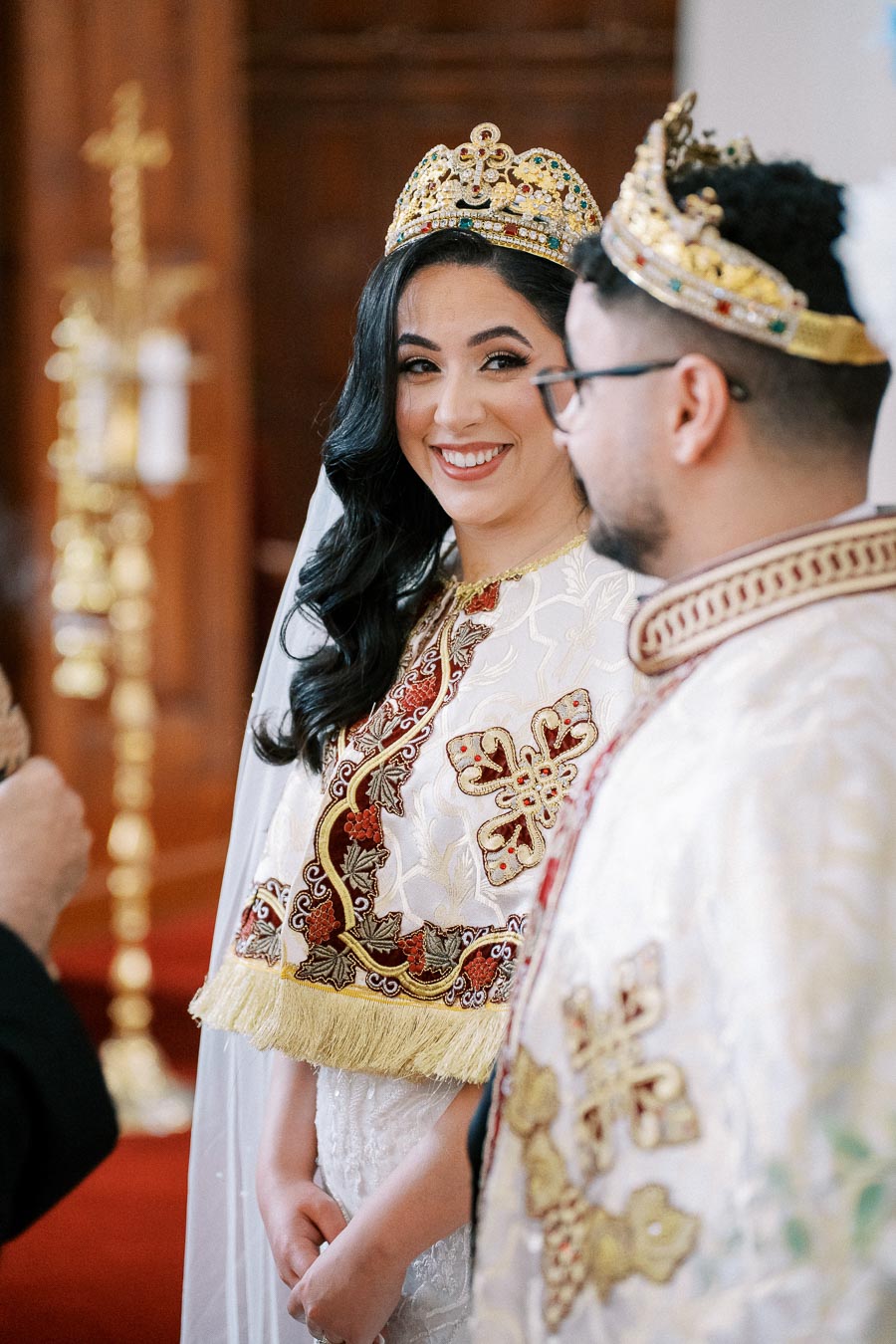Bride and groom in traditional ornate robes and crowns smiling during a wedding ceremony in a church.