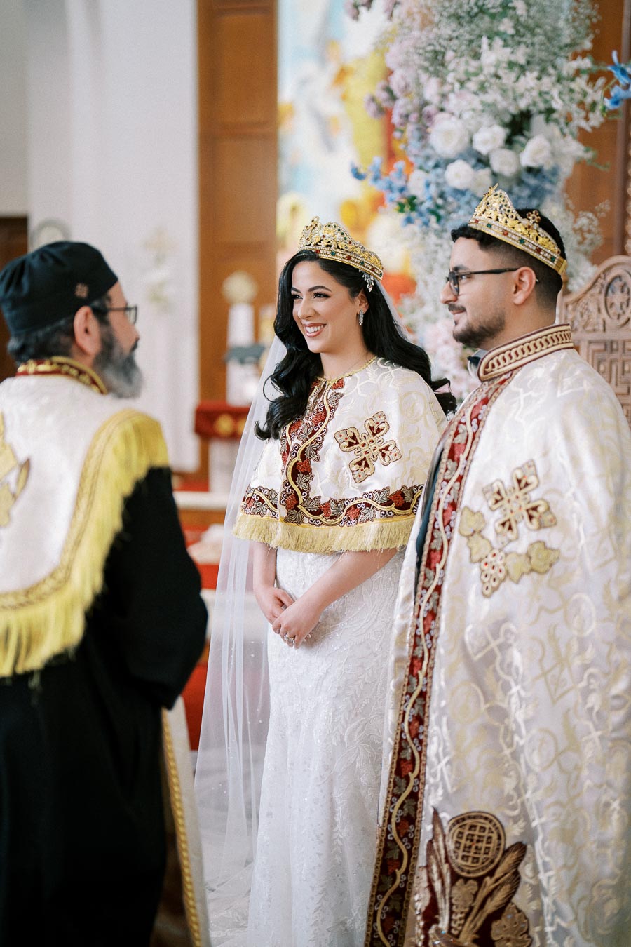A couple wearing ornate crowns and traditional robes stands before a priest during a religious wedding ceremony, surrounded by floral decorations in a beautifully decorated church setting.