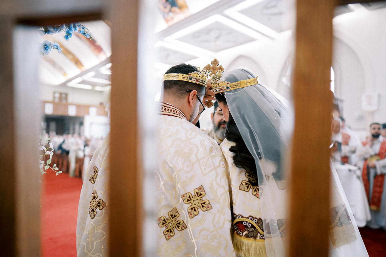 A couple wearing ornate crowns during an Orthodox wedding ceremony, with a view of the church interior and attendees in the background.