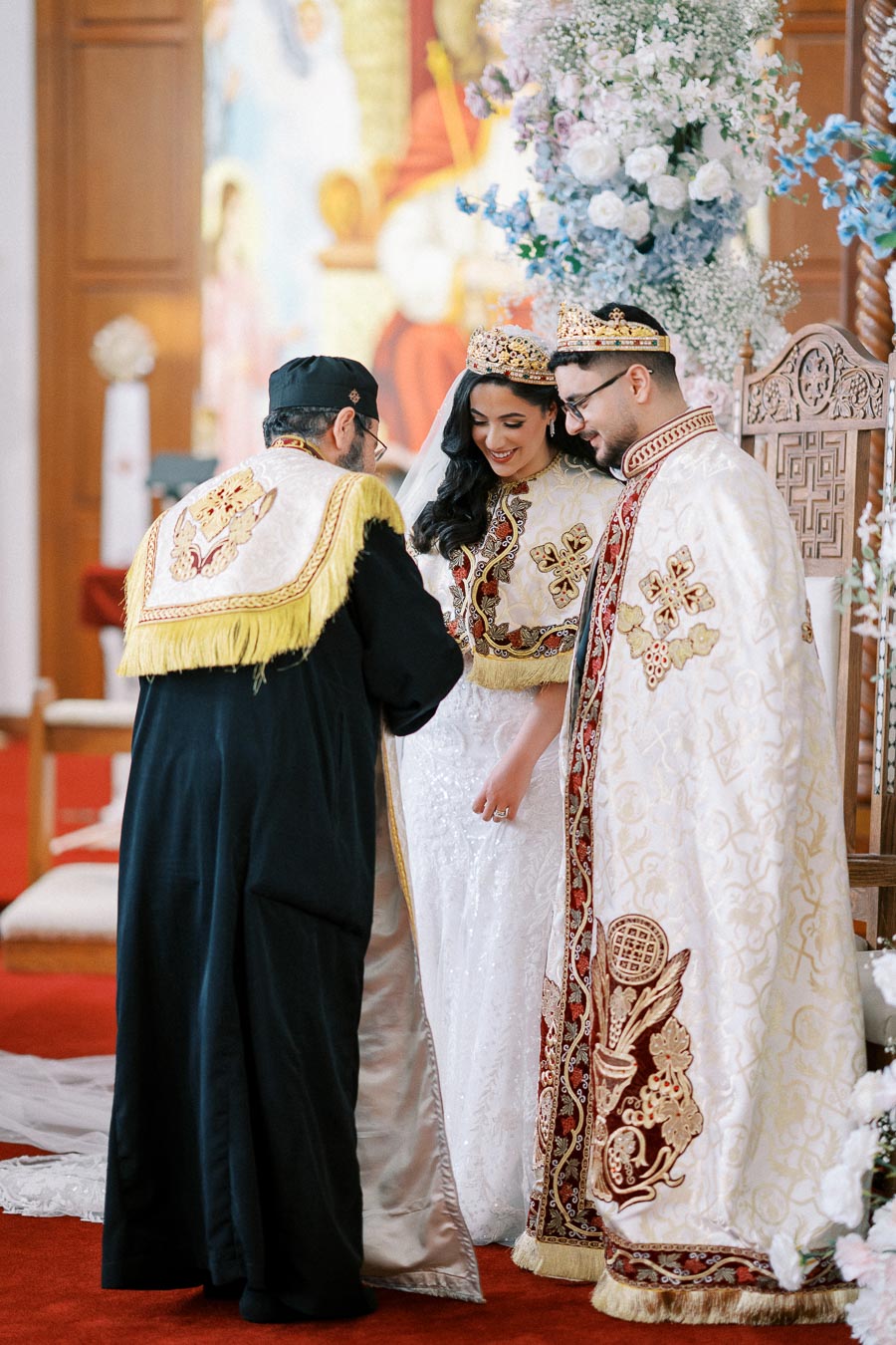 Orthodox wedding ceremony with bride and groom wearing traditional embroidered attire and crowns, standing before a priest in a richly decorated church setting.