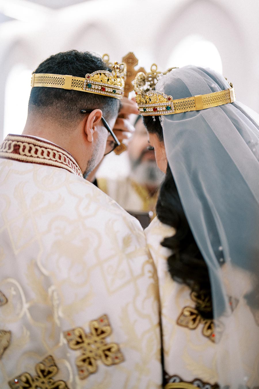 A bride and groom in traditional ceremonial attire wearing ornate golden crowns during a wedding service.