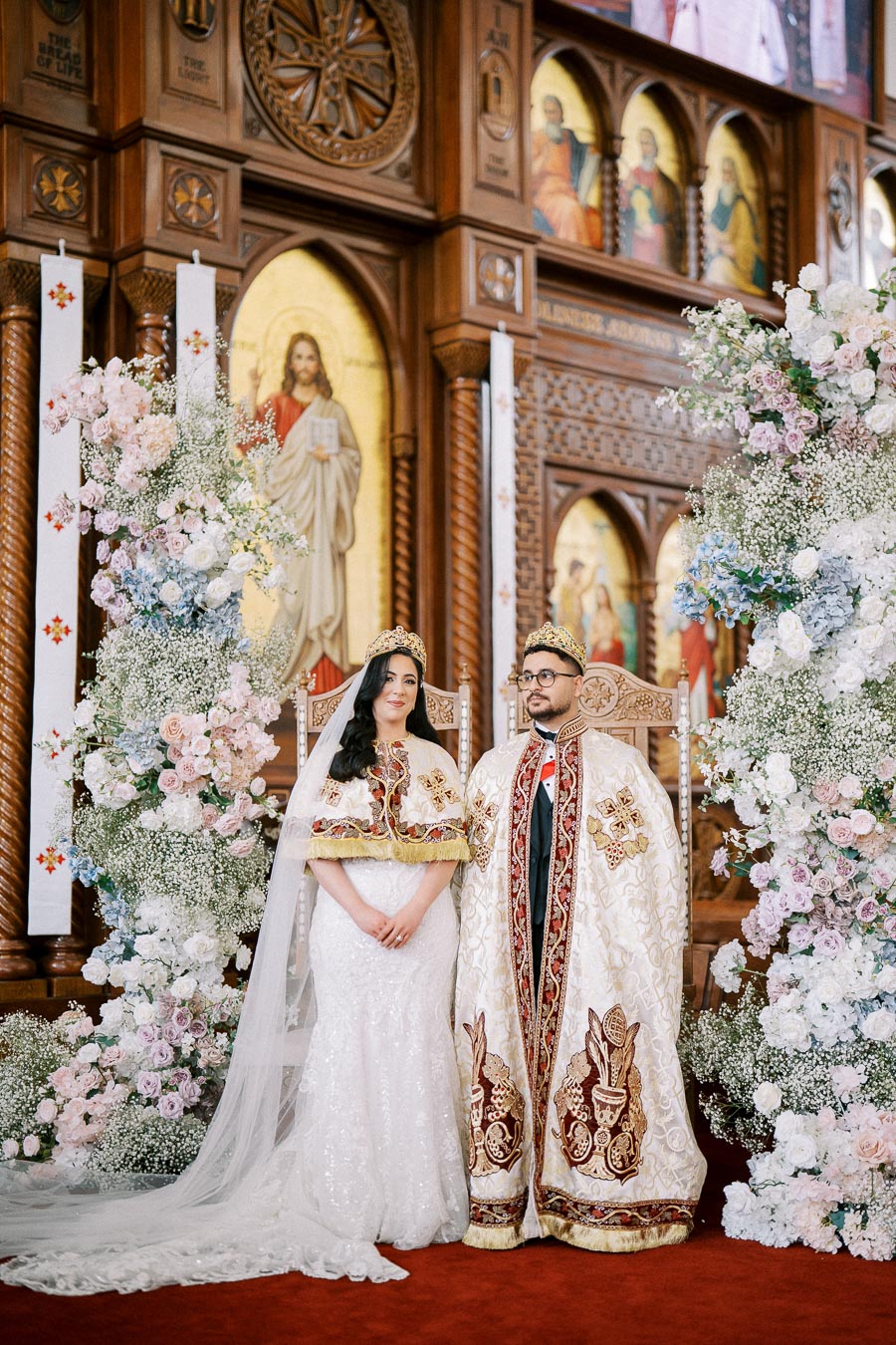 A bride and groom in traditional attire stand together in an ornate church setting, surrounded by elaborate floral arrangements and religious iconography. The couple wears crowns and ceremonial robes, highlighting a cultural or religious wedding ceremony.