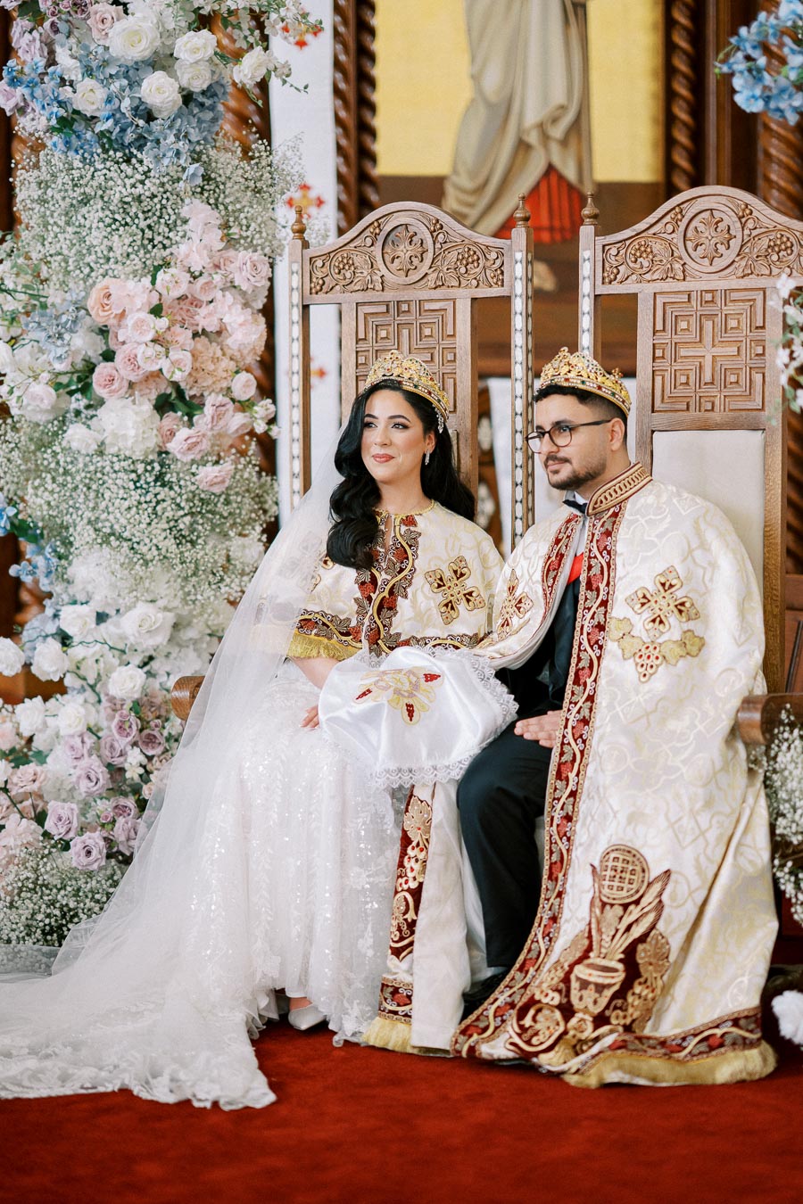 A bride and groom in ornate traditional attire sit on intricately carved chairs, surrounded by elegant floral arrangements during a wedding ceremony.