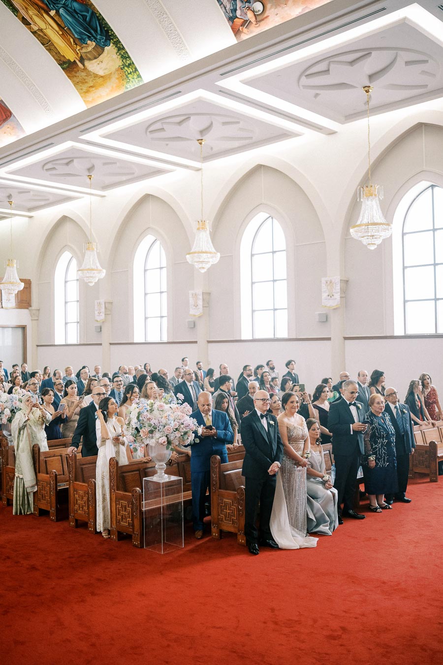 A large gathering of well-dressed guests attends a formal ceremony inside an ornate church with high ceilings and elegant chandeliers, seated on wooden pews with floral arrangements.