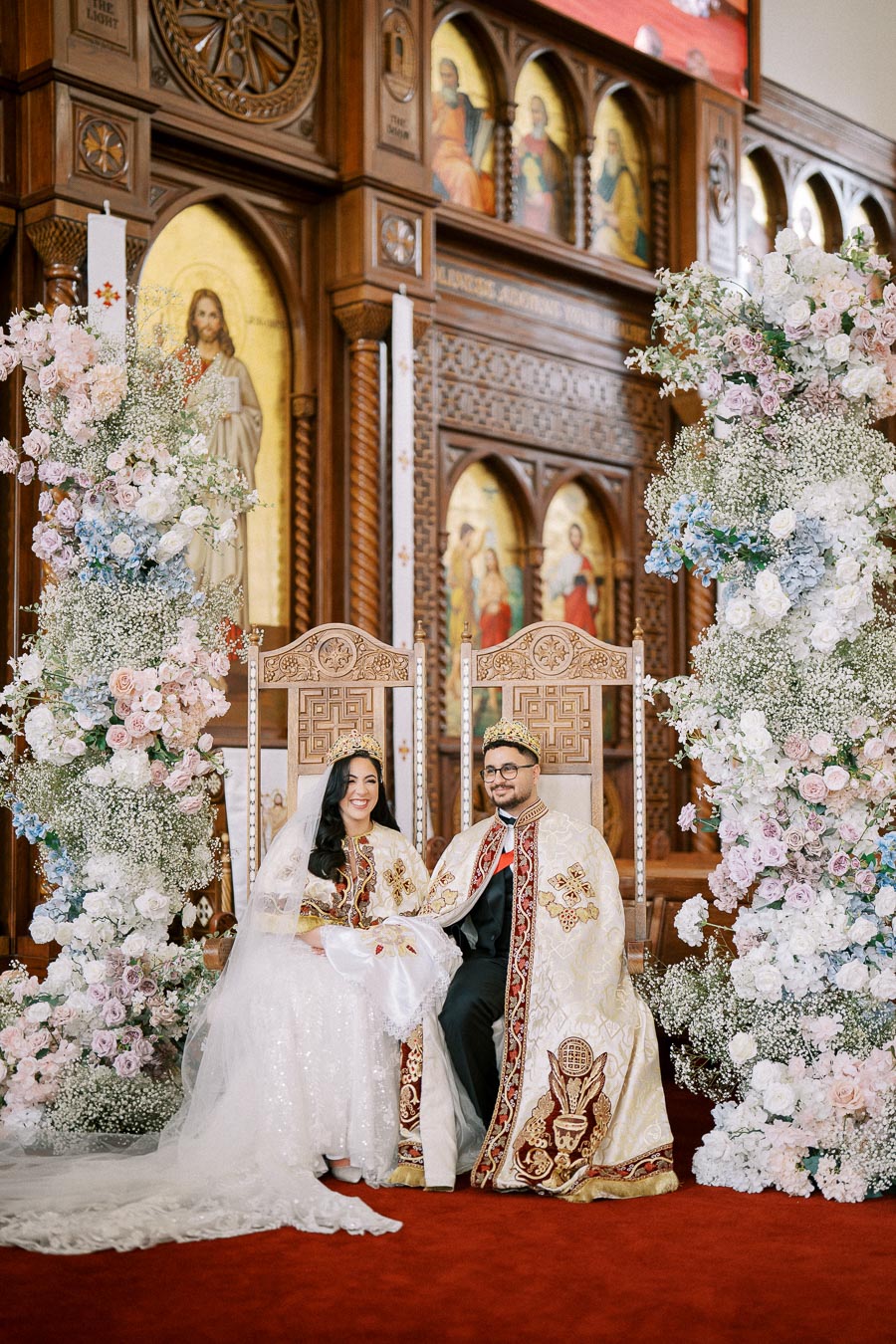 Alt text: Bride and groom wearing traditional ornate robes and crowns sitting on decorative chairs in a beautifully adorned church, surrounded by elegant floral arrangements and intricate wooden carvings.