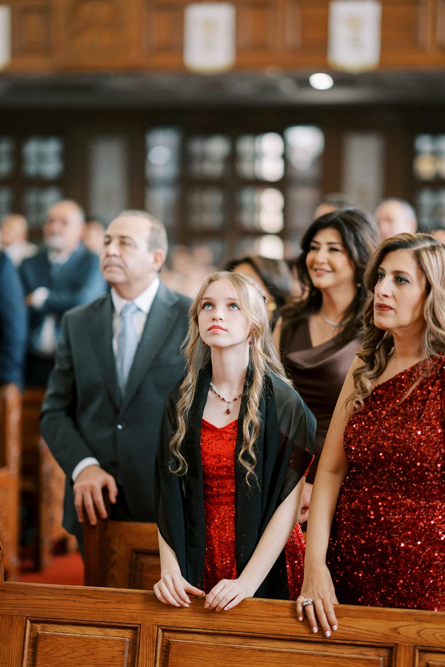 A group of people attentively standing during a formal event in a church, with a young girl in a red dress looking up, surrounded by adults in elegant attire.