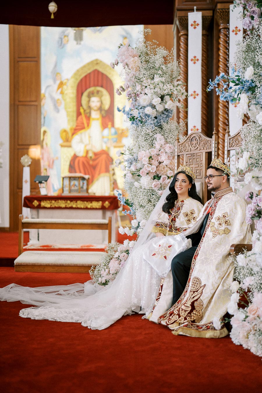 A bride and groom dressed in traditional ornate robes and crowns sit on a decorated throne during a vibrant and colorful Christian wedding ceremony, surrounded by lush floral arrangements inside a church.