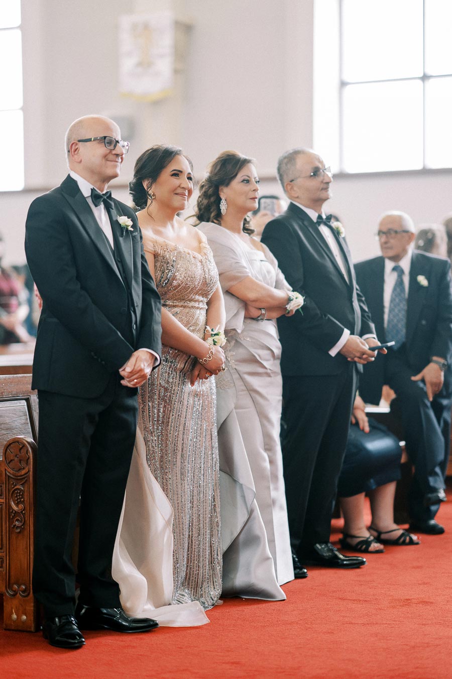 A group of well-dressed individuals stands inside a church, dressed in formal attire. Two men in black tuxedos and two women in elegant gowns, smiling as they look towards the front. The church interior is softly lit, featuring wooden pews and large windows.