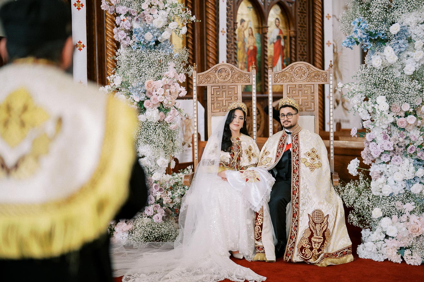 A bride and groom dressed in ornate traditional garments and crowns sit on intricately carved chairs during a religious wedding ceremony, surrounded by floral arrangements with pink and white roses in a beautifully decorated church.
