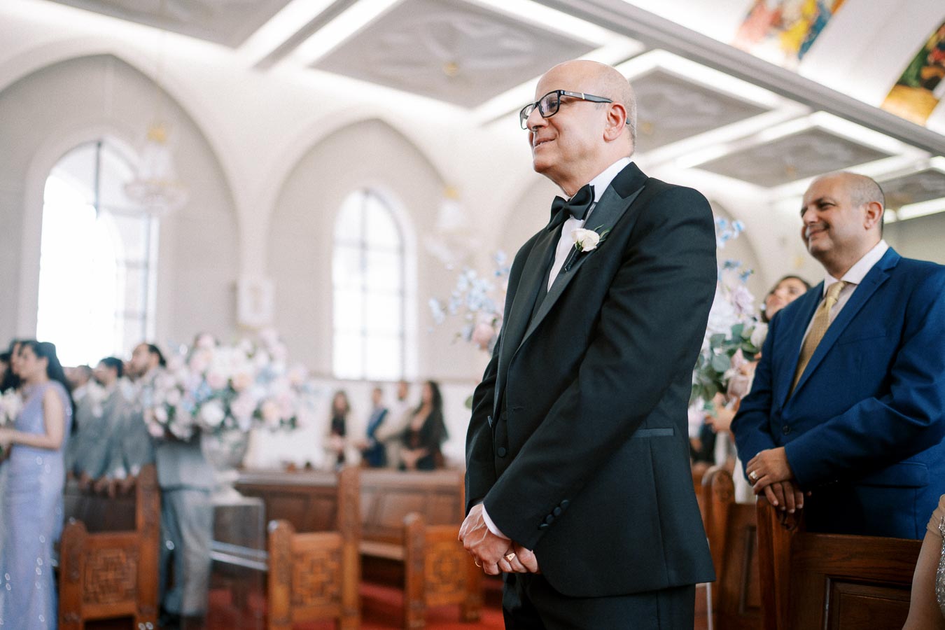 A smiling man in a suit stands in a beautifully decorated church, surrounded by guests during a wedding ceremony.
