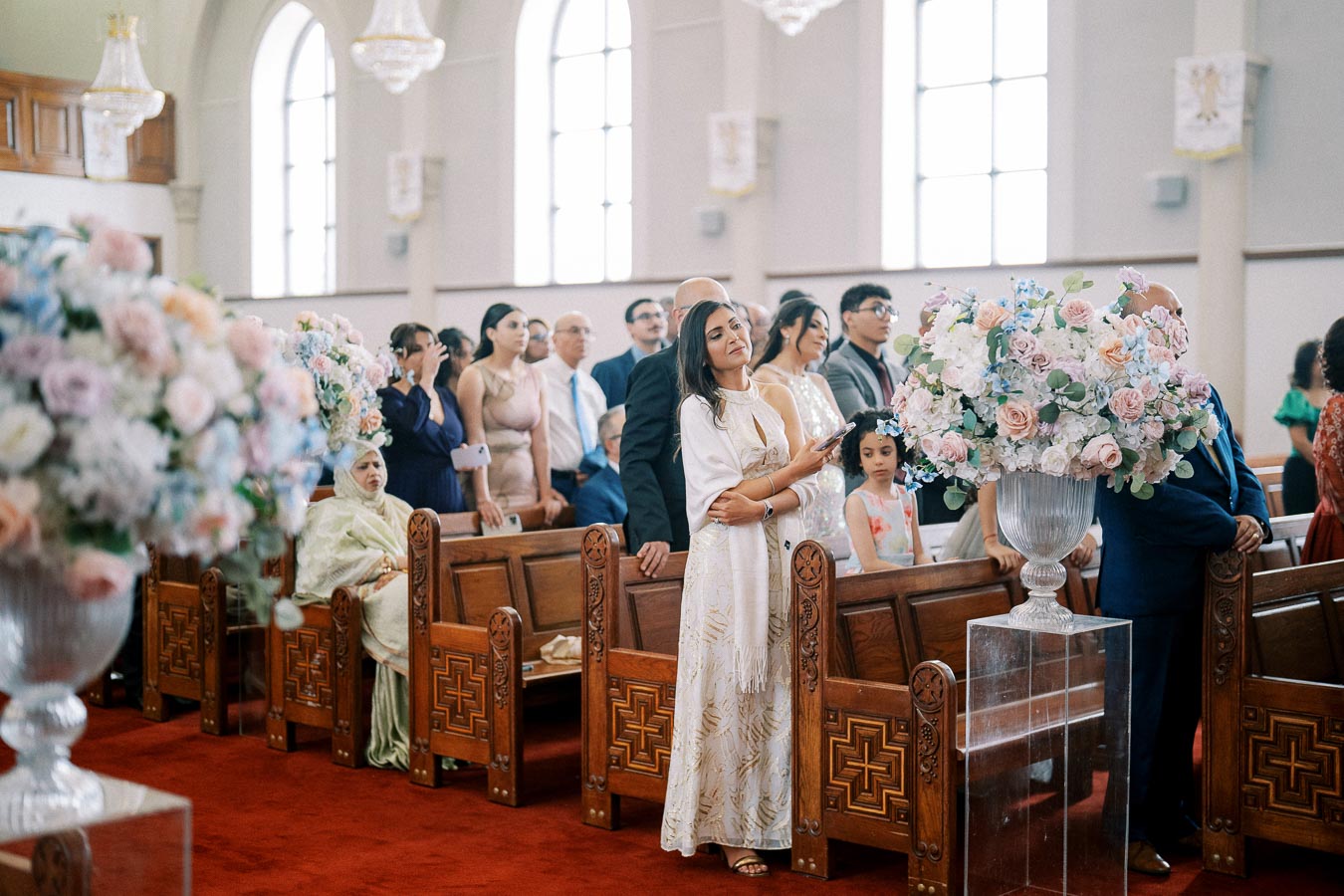 A diverse group of people stands inside a beautifully decorated church, attending a wedding ceremony. Large floral arrangements and elegant chandeliers accentuate the serene atmosphere, as light streams through tall arched windows, illuminating the ornate wooden pews.