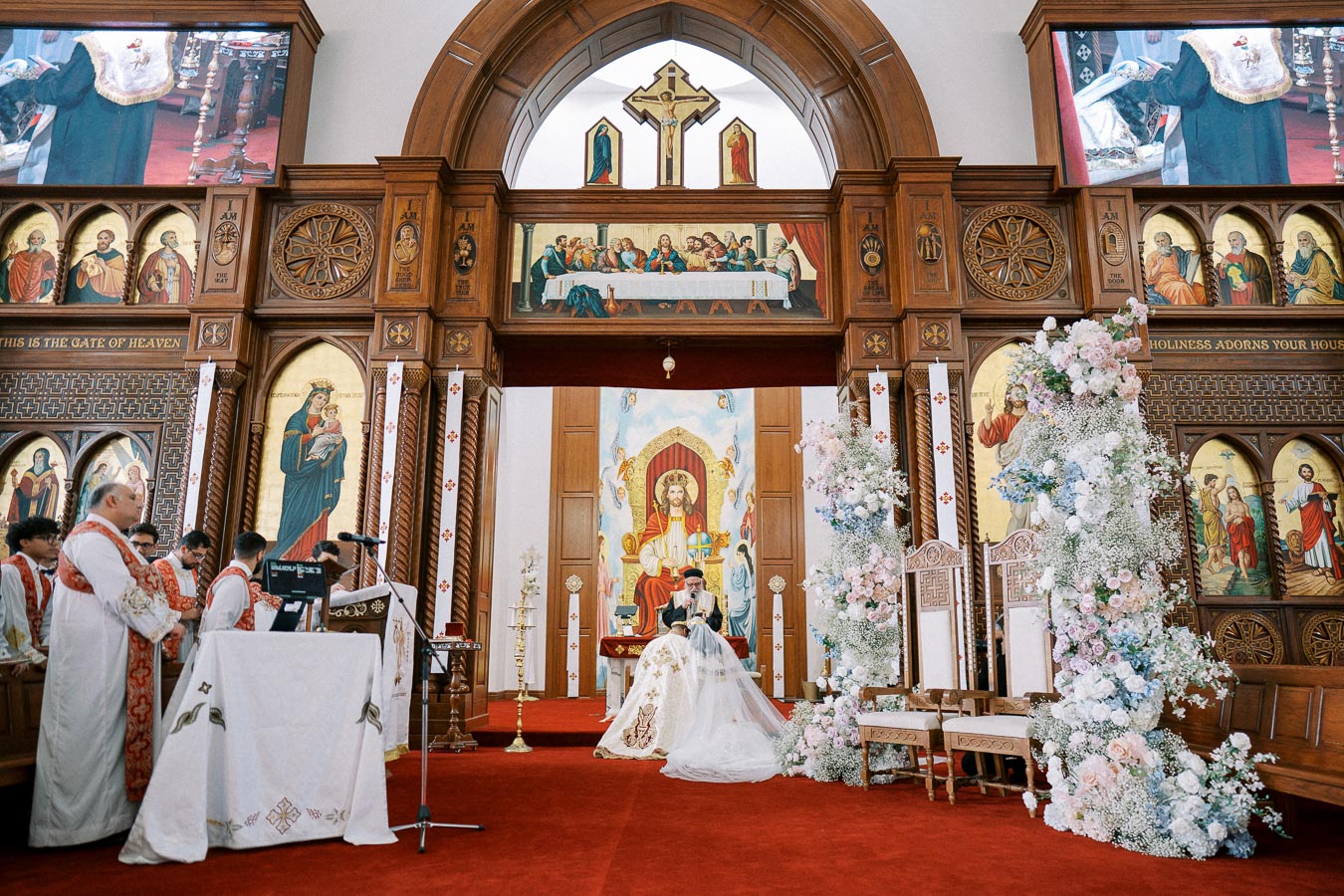 Interior of a beautifully decorated church with ornate wooden fixtures, religious icons, and a floral arrangement surrounding chairs. Priests in white and red vestments stand near the altar during a ceremonial event.