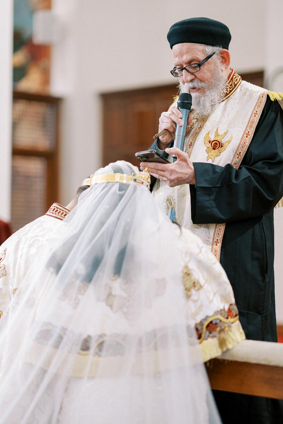 Priest conducting a traditional Coptic wedding ceremony, reading from a script while a bride in a veiled gown kneels before him in a richly decorated church setting.