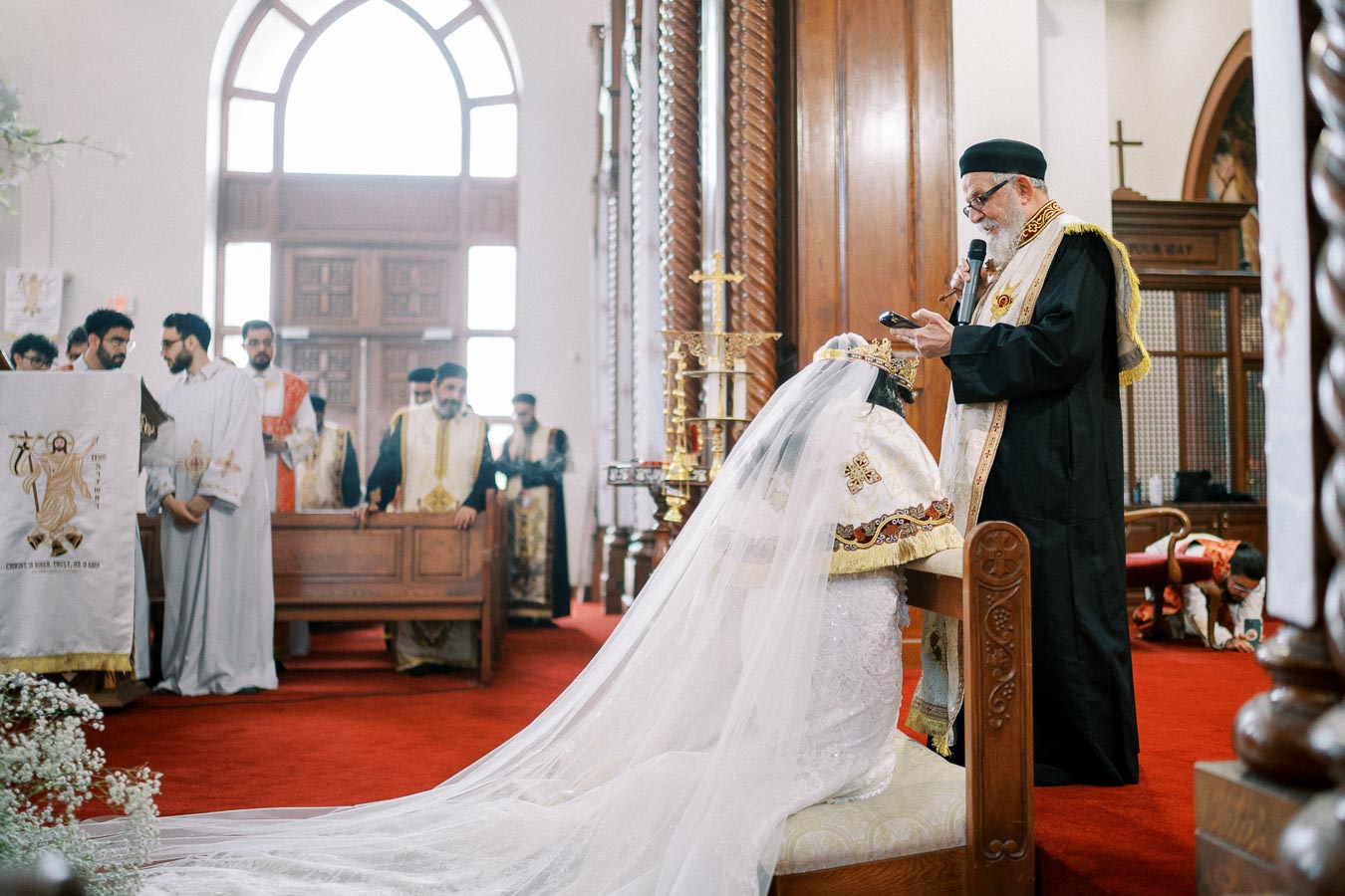 Orthodox priest officiating a wedding ceremony inside a beautifully adorned church, with a bride wearing an ornate veil and crown, surrounded by clergy and decorative religious symbols.