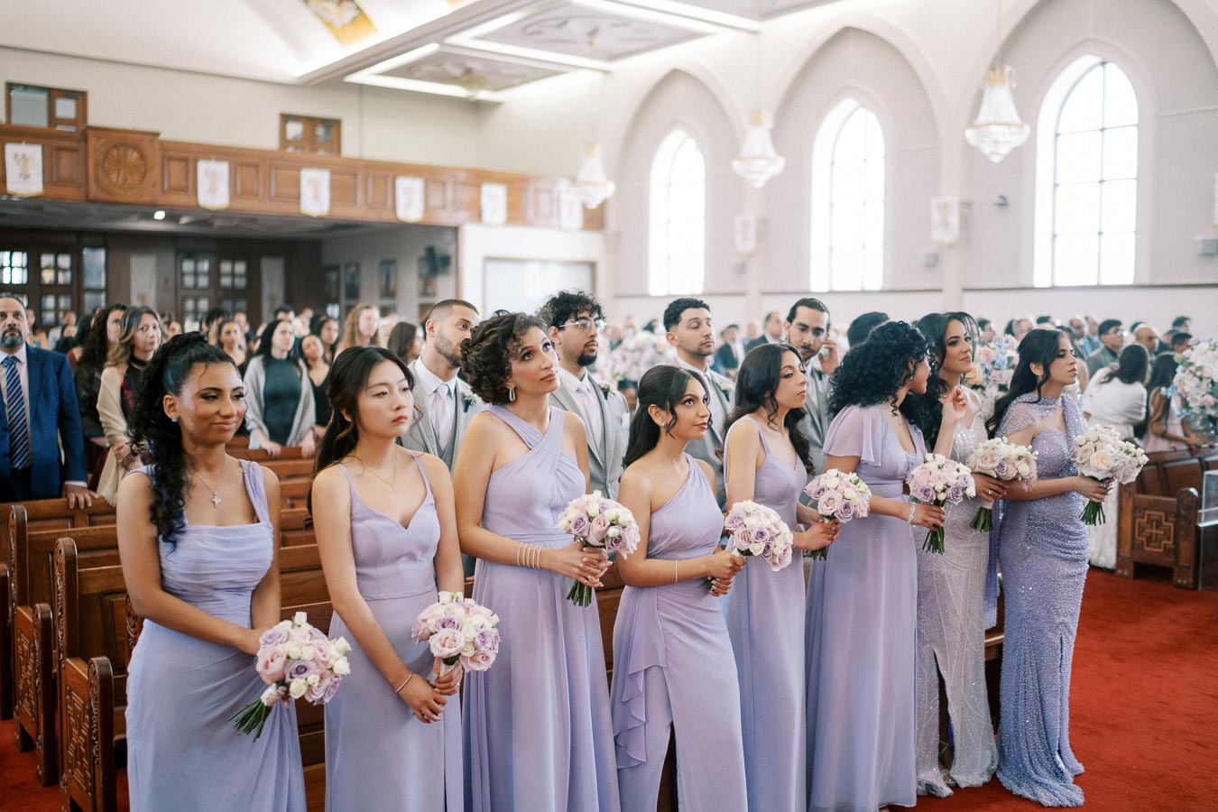 Bridesmaids in lavender dresses holding bouquets stand in a church with arched windows, surrounded by guests during a wedding ceremony.