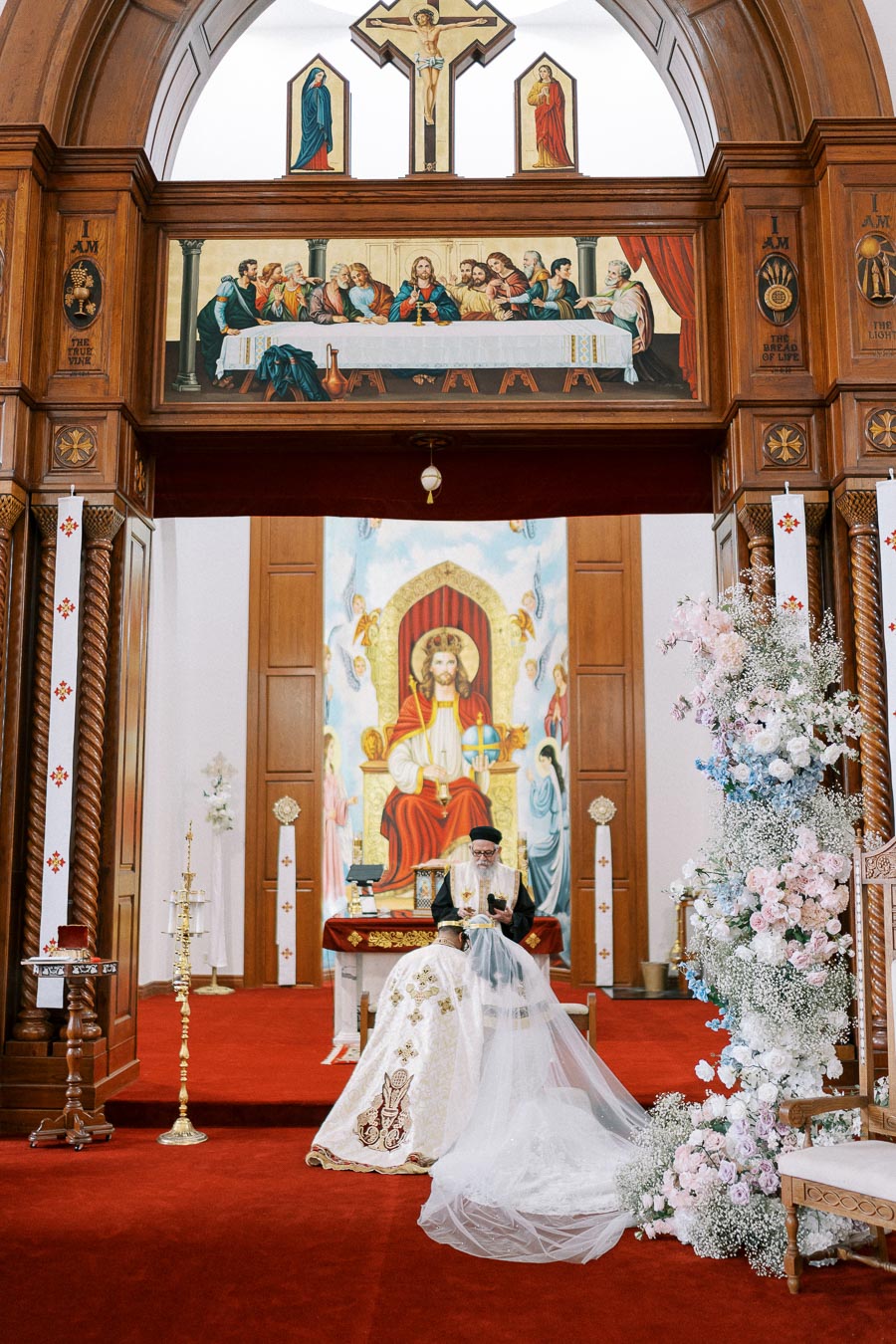 Alt text: A beautifully decorated church interior featuring ornate religious artwork and floral arrangements. A priest in ceremonial attire is seated in front of a large painting depicting a religious scene. The altar is adorned with intricate designs, and the atmosphere is reverent and serene.