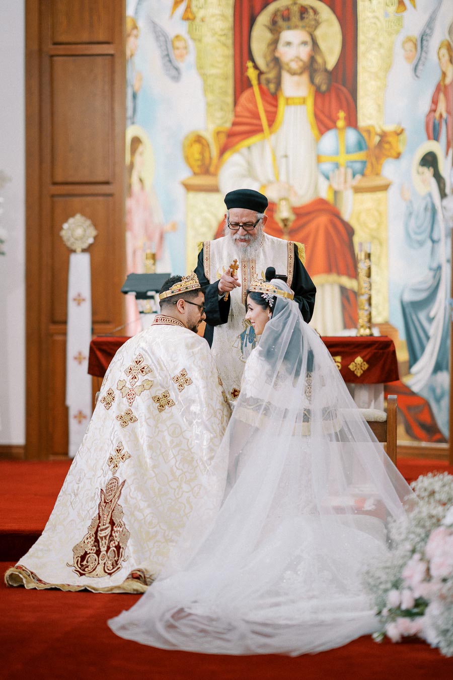 Orthodox wedding ceremony with priest blessing bride and groom in a church adorned with religious icons and floral decorations.