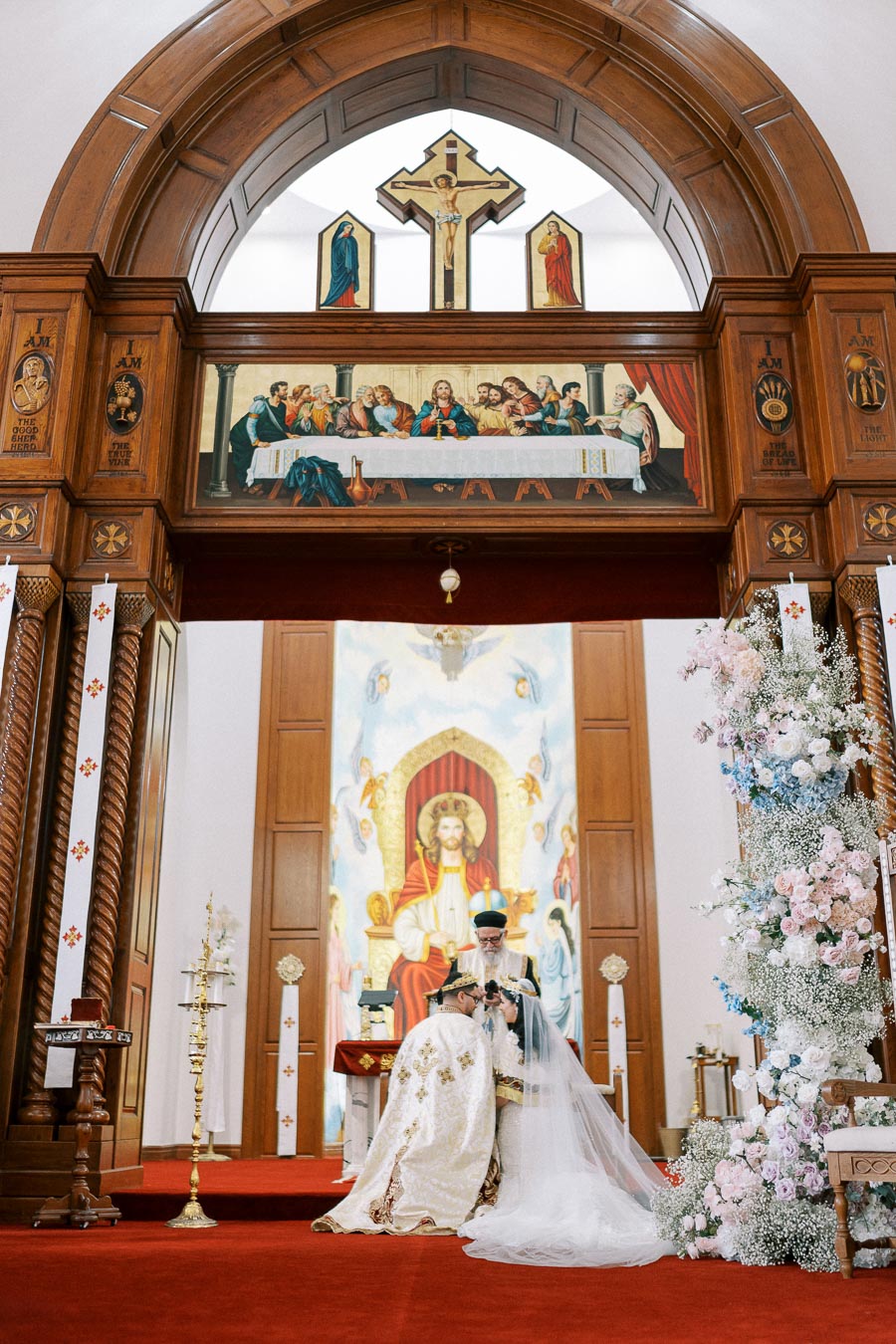 A bride and groom kneel before a priest during a traditional church wedding ceremony, surrounded by ornate religious artwork and floral arrangements.