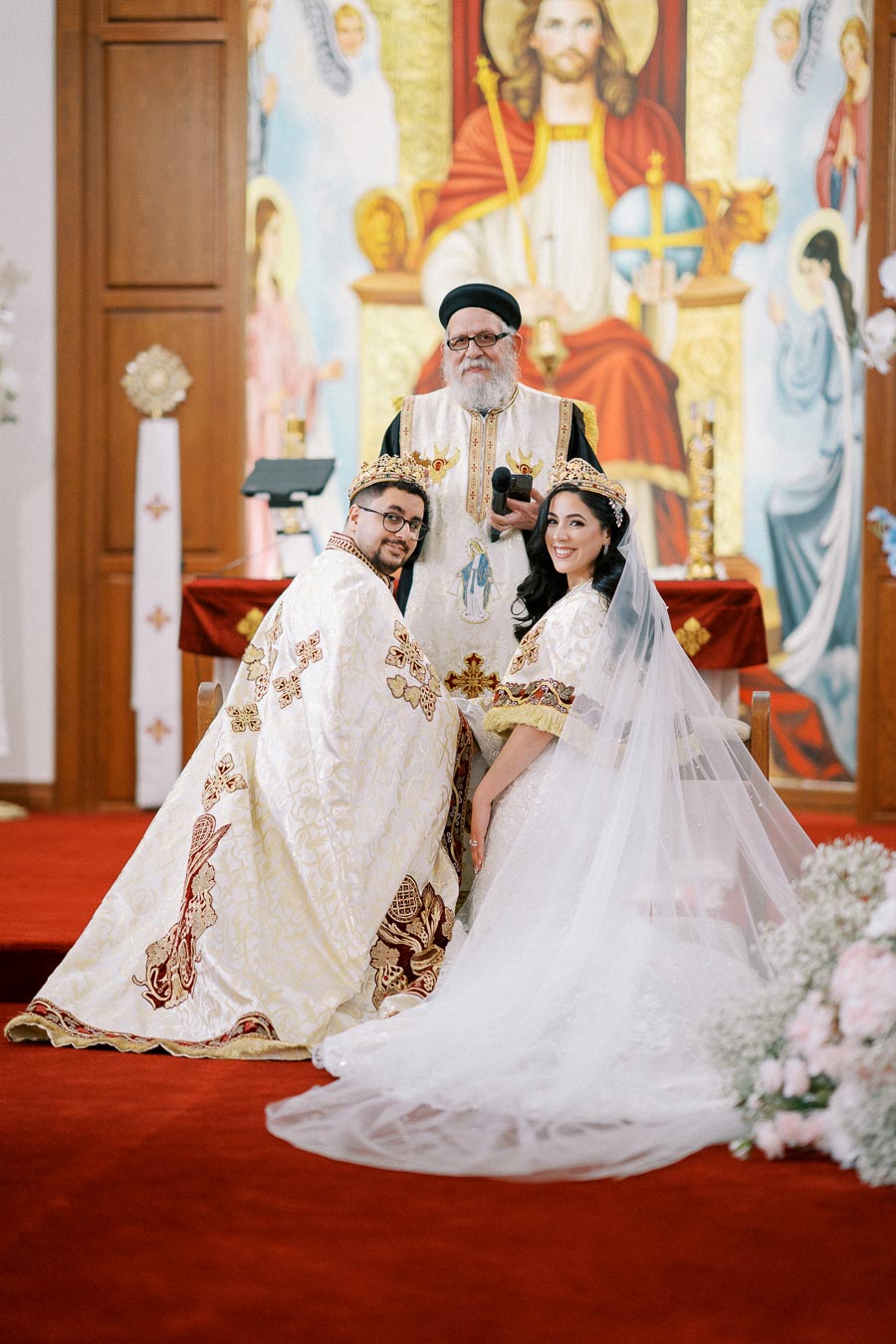 Orthodox wedding ceremony with bride and groom wearing ornate crowns, standing before a priest in a church adorned with religious artwork.