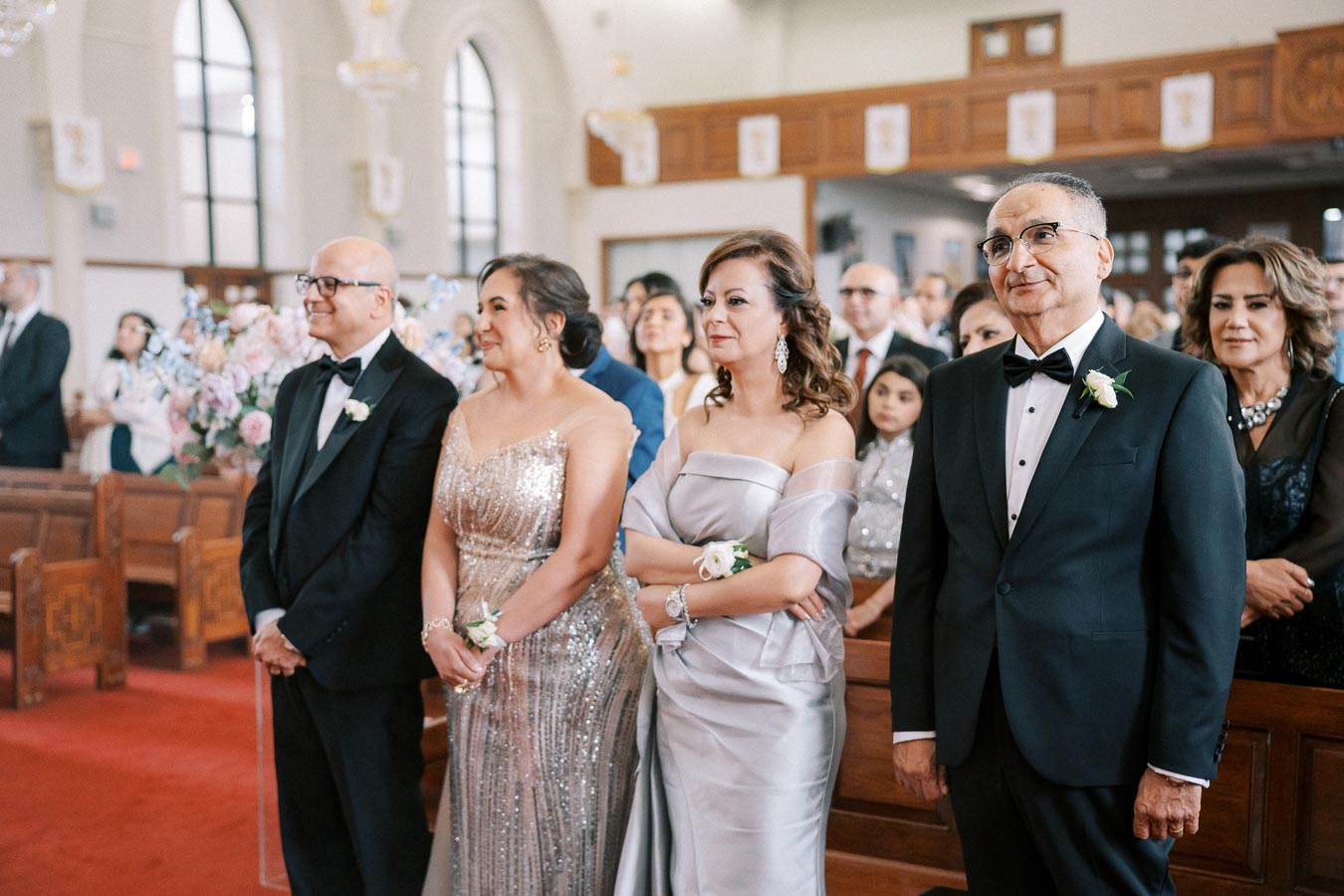 A group of elegantly dressed wedding guests standing in a church with wood and floral decorations, showcasing formal attire including suits and sparkling gowns.