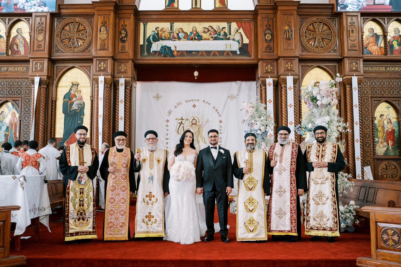 A bride and groom stand together between priests in traditional religious attire inside a richly decorated church with ornate wooden panels and religious iconography.