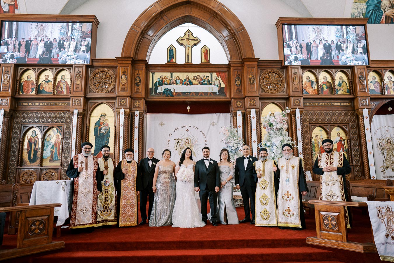 A wedding ceremony in a beautifully decorated church, with a bride and groom surrounded by family and clergy members in traditional attire, standing in front of an ornately designed altar featuring religious iconography and floral arrangements.