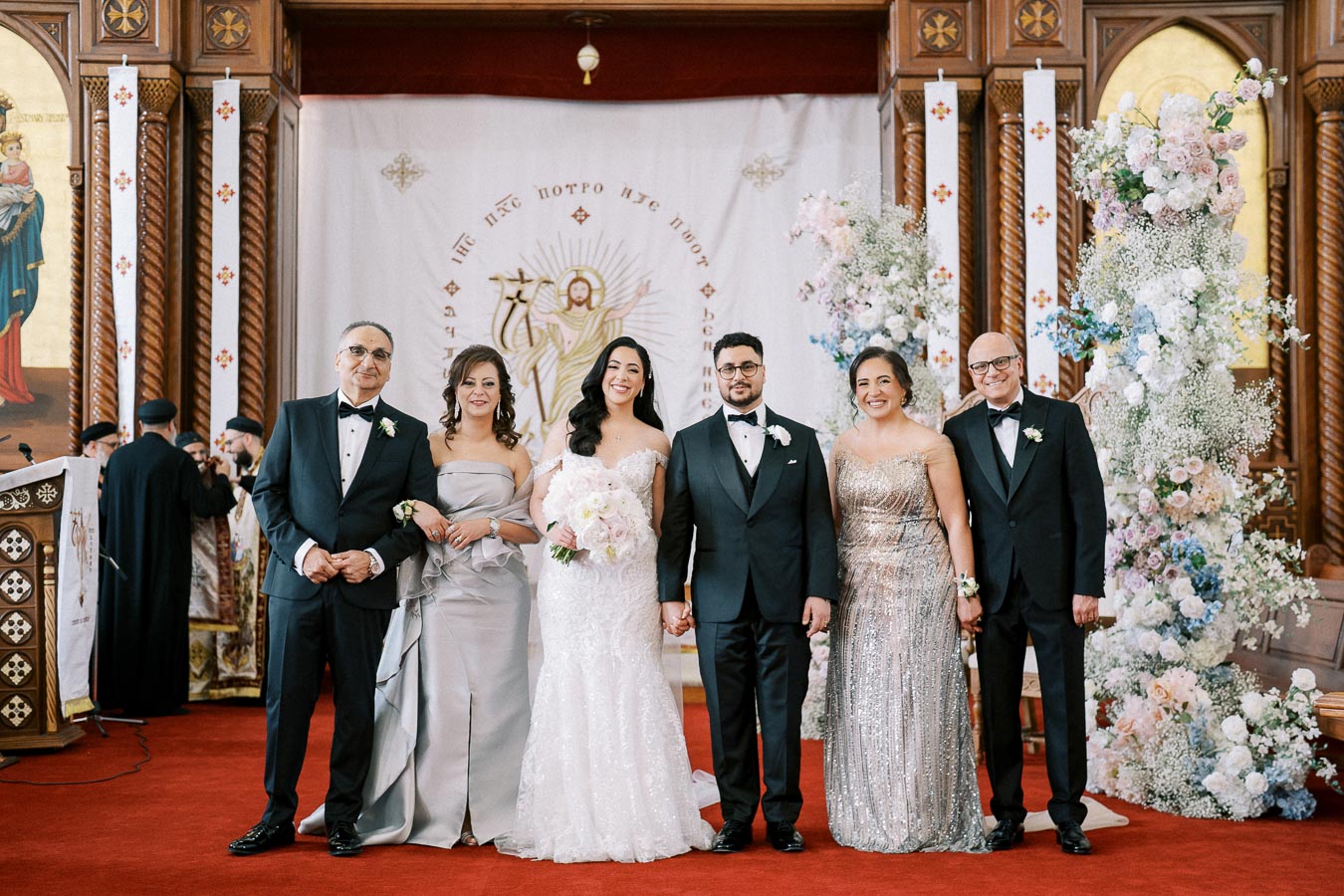 A bride and groom standing with four elegantly dressed guests in a beautifully decorated church interior, featuring floral arrangements and intricate wooden designs, during a wedding ceremony.