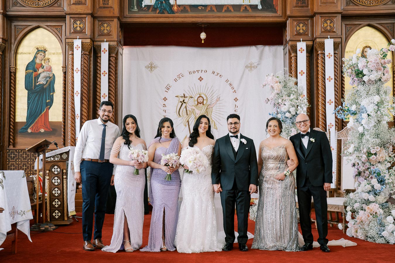 A group of six people posing in elegant attire inside a richly decorated church with religious icons and floral arrangements, likely celebrating a special occasion or ceremony.
